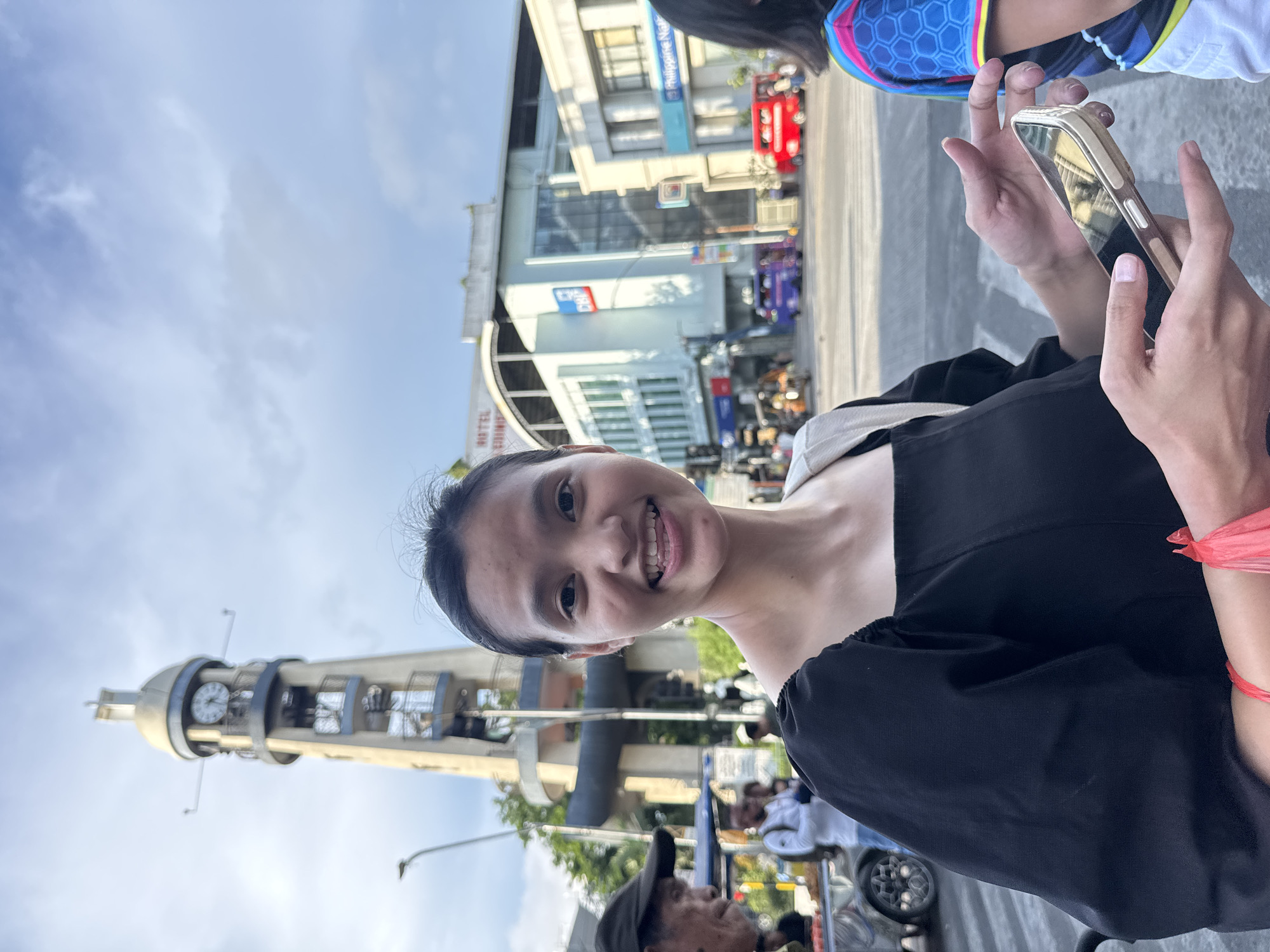 This media file depicts a street scene in Davao City, Philippines. A young woman in the foreground is smiling at the camera. She is wearing a black top with a white trim around the neckline and what appears to be black shorts. Her right hand is visible holding a gold-colored smartphone. Behind her, a distinctive clock tower, a landmark in Davao City, is visible against a bright, cloudy sky, suggesting it is daytime. The background shows urban elements, including buildings with signage, traffic, and other people, some of whom are wearing athletic attire. One building has a sign that reads "Philippine National Bank". The overall atmosphere appears casual and public, possibly indicating a day out or an event.