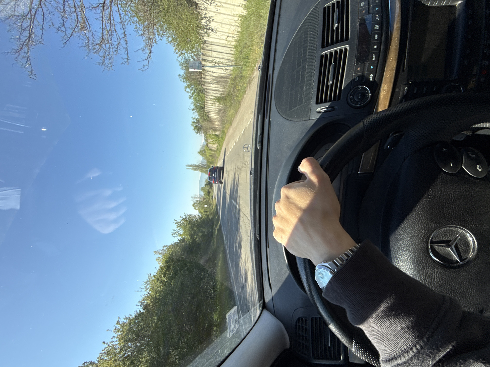 A person is driving a Mercedes-Benz on a road in Buhovo, Bulgaria. The person's left hand is visible holding the steering wheel. The dashboard and interior of the car are visible. Outside the car, the road stretches ahead with greenery on either side and a vehicle in the distance. The sky is blue with tree branch reflections visible in the windshield. A concrete wall runs parallel to the road on the right.