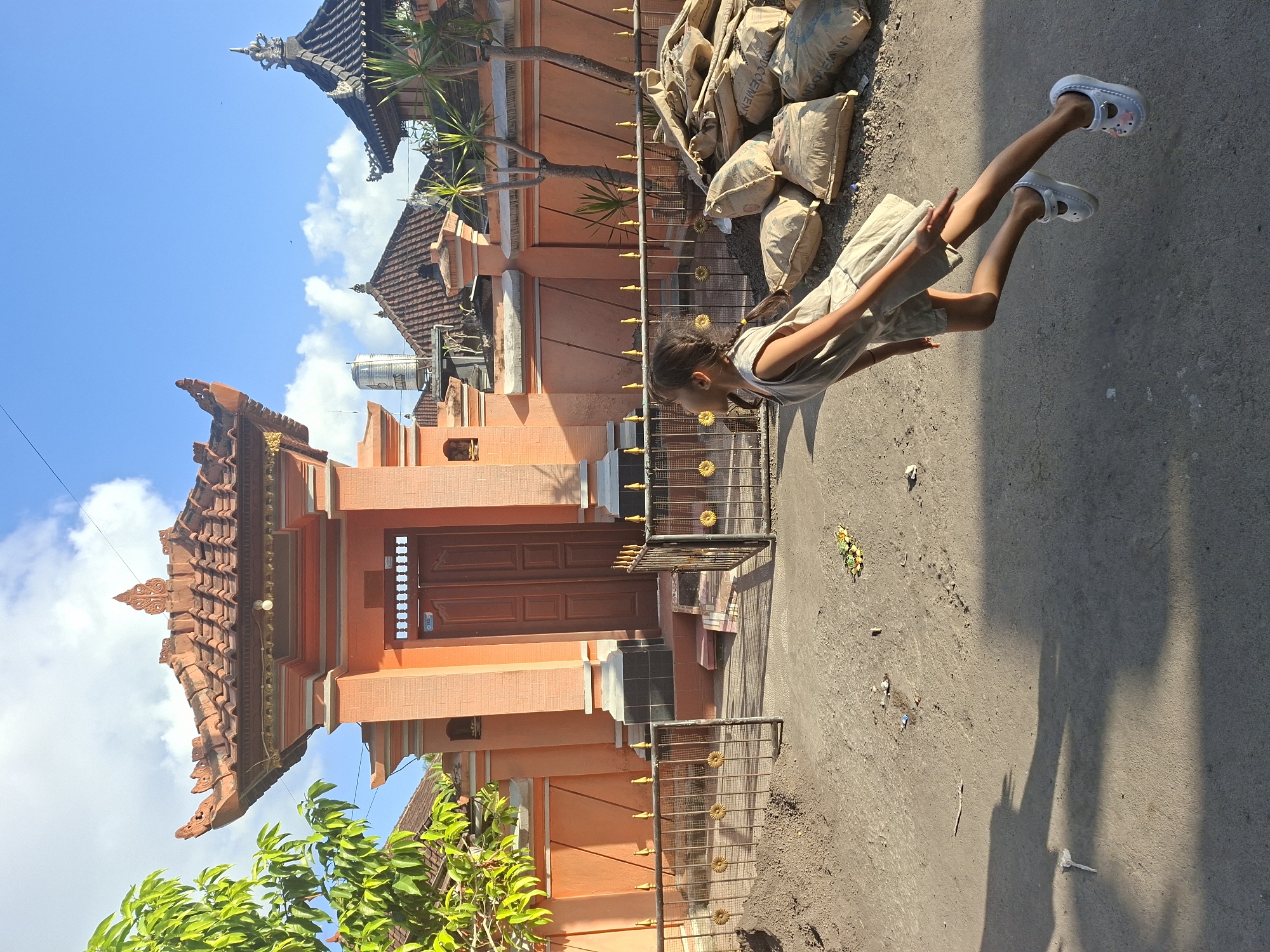 A young girl with braided hair, wearing a light-colored dress and white shoes, is captured mid-air in a dynamic pose, appearing to be jumping or floating. She is positioned on a dirt road in front of a coral-colored building with an ornate tiled roof. The building features a large wooden double door and a decorative fence with golden embellishments. To the right of the girl, a pile of sandbags is stacked. The background shows more buildings with similar architectural styles, a clear blue sky with scattered clouds, and greenery from a nearby tree. The sun casts a long shadow of the girl onto the ground.