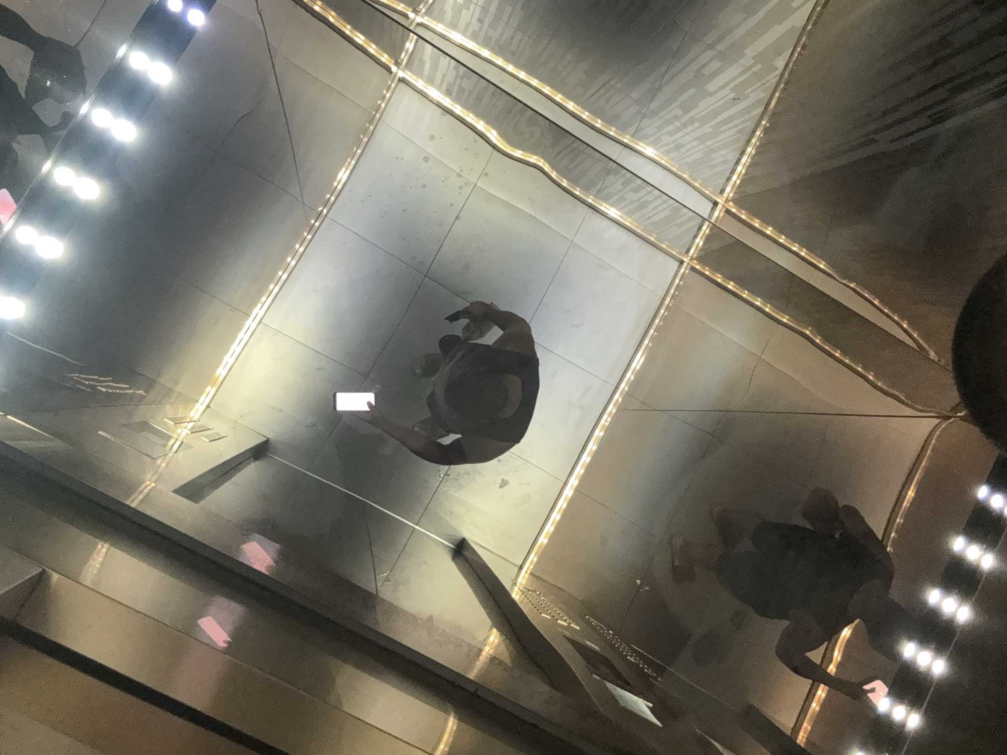An overhead shot captures a modern, highly reflective indoor space, possibly an elevator or a corridor, in Kuala Lumpur, Malaysia. The main subject, a person, is seen from above, standing near the center of the frame, looking down at a smartphone with a white case held in their hands. The person's form, along with faint reflections of other elements, is cast upon the glossy, dark-toned floor and wall surfaces.
The environment is characterized by prominent linear light strips embedded into the ceiling or walls, forming a geometric, grid-like pattern that reflects brightly off the polished surfaces, creating a sense of depth and futuristic aesthetics. These lights are a significant source of illumination, contributing to the strong reflections and high contrast in the scene. The surfaces appear to be made of polished stone, metal, or mirrored panels, showing some subtle smudges or patterns.
Another faint, darker reflection of a person is visible towards the right side of the frame, partially obscured, suggesting the presence of at least one other individual or another reflection of the main subject. The overall impression is one of a sleek, contemporary interior, illuminated by artificial light, with a focus on reflections and geometric lines. There is no visible text in the image.
