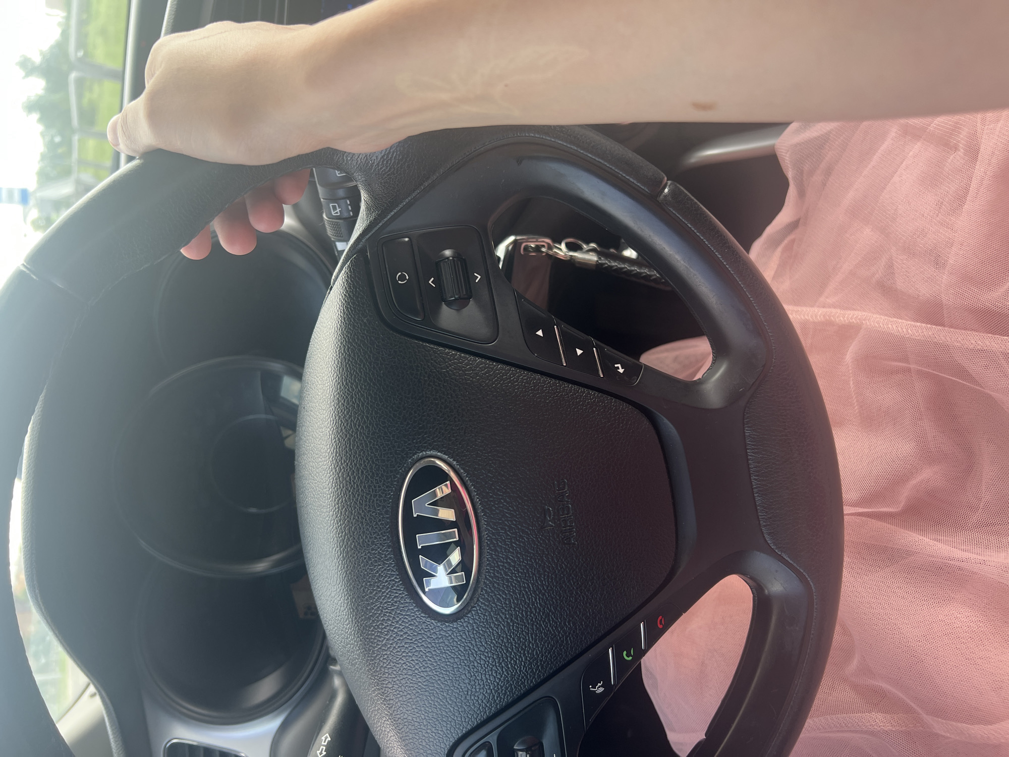 A person's hands are visible on the steering wheel of a Kia car. The steering wheel is black with the Kia logo in the center. Various buttons and controls are present on the steering wheel. A set of keys with a keychain is hanging from the steering column. The interior of the car is dimly lit. Outside the car, greenery and a fence are visible through the windshield, suggesting an outdoor environment, possibly a road. The image is taken from a close-up perspective, focusing on the steering wheel and the driver's hands. There is no discernible information about the specific location within Magnitogorsk, Russia, or any indication of actions or interactions beyond the implied act of driving.