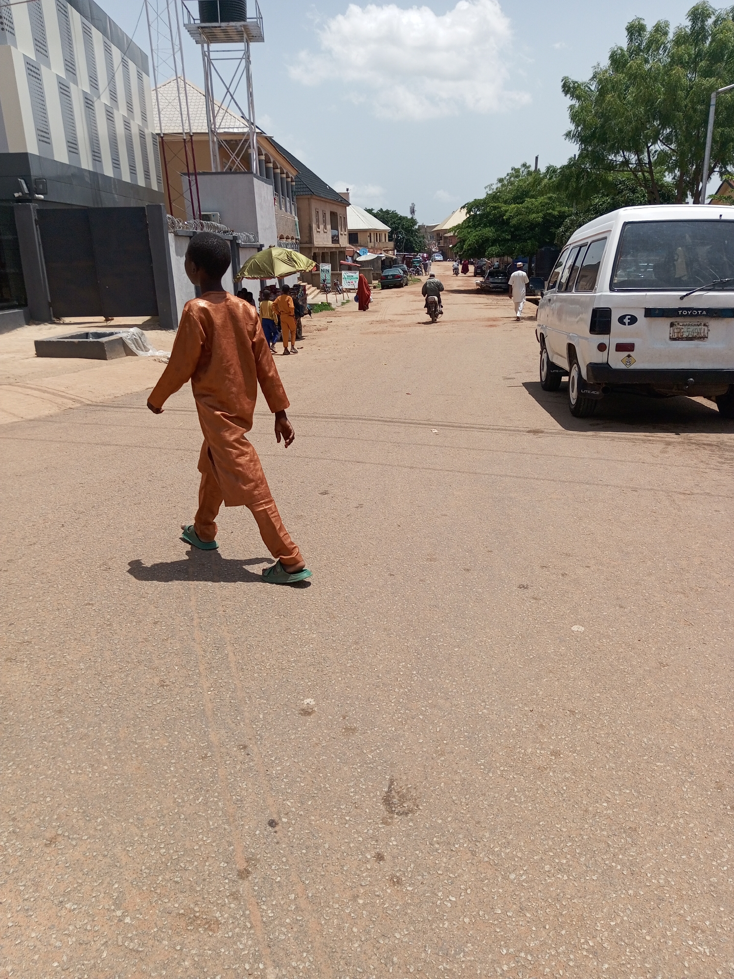 This is a mid-day, eye-level, outdoor, full-shot photograph taken on a paved road in Bukuru, Nigeria. The scene is dominated by a young boy walking towards the left of the frame, wearing an orange traditional outfit and green sandals. He casts a shadow on the asphalt road, which is speckled with small stones. In the background, a white Toyota van is parked on the right side of the road. Further back, several people are visible, including individuals on a motorcycle and others walking, some beneath a yellow umbrella. Buildings line both sides of the street, featuring modern architectural elements like glass facades and traditional structures with peaked roofs. Green trees are scattered throughout the background, and the sky is partly cloudy. The overall atmosphere suggests a bustling urban environment under a bright sky.