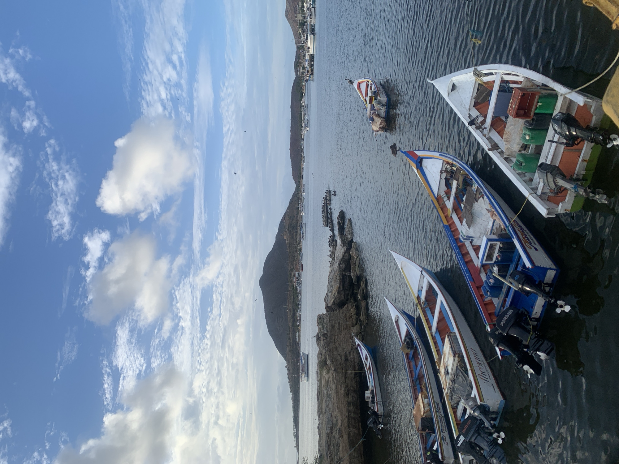 The image depicts a tranquil scene of a harbor in Juan Griego, Venezuela, bathed in the soft light of early morning or late afternoon. The sky is a canvas of pale blue, adorned with scattered white clouds that catch the sunlight, with a few distant birds soaring high above. In the foreground, the calm, dark water of the bay reflects the muted colors of the surrounding environment.
Several fishing boats are moored in the harbor. In the immediate foreground, three narrow, colorful boats are lined up. The closest boat is white with red and green trim along its hull, and an orange-colored interior. It has two outboard motors mounted on the stern and contains green containers and a red wooden crate. Next to it, another boat is white with blue and red trim, also featuring two outboard motors. This boat has a blue interior and a name visible on its side which reads "Shoeiva". A third boat, further back and slightly out of focus, shares a similar design with blue and red trim.
In the middle ground, a more distant, larger boat with a white cabin and a dark hull sails across the water. To the left of this boat, a rocky outcrop juts into the water, with numerous seabirds perched on its surface. Further out in the bay, a collection of other boats, including larger vessels and what appear to be sailboats, are anchored.
The background is dominated by a large, dark, tree-covered mountain that rises majestically from the coastline. A small town with buildings and docks is visible nestled at the base of the mountain and along the shoreline. The overall atmosphere is one of peaceful maritime activity, characteristic of a coastal Venezuelan town.