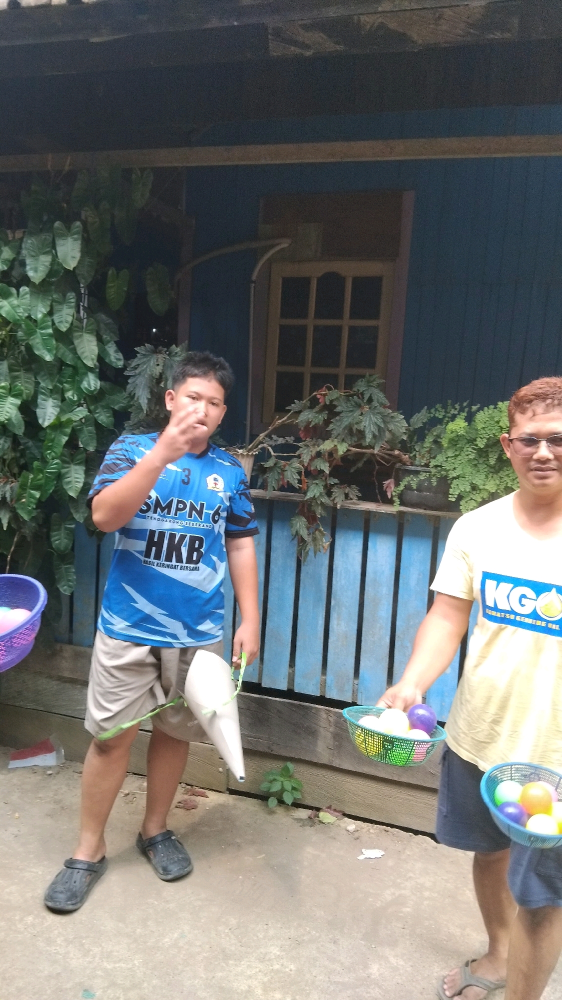 In Bangun Rejo, Indonesia, two individuals stand outside a blue wooden building. On the left, a young boy wearing a blue t-shirt with "SMPN-6" and "HKB" printed on it, and light-colored shorts, stands near a purple basket containing colorful balls. He is holding a white cone-shaped object with green string. To his right, a man wearing a yellow t-shirt with "KG0" printed on it, and dark shorts, holds two baskets filled with colorful balls. The background features lush green foliage and a blue wooden fence. The ground is dirt and concrete. The scene appears to be a casual outdoor gathering.