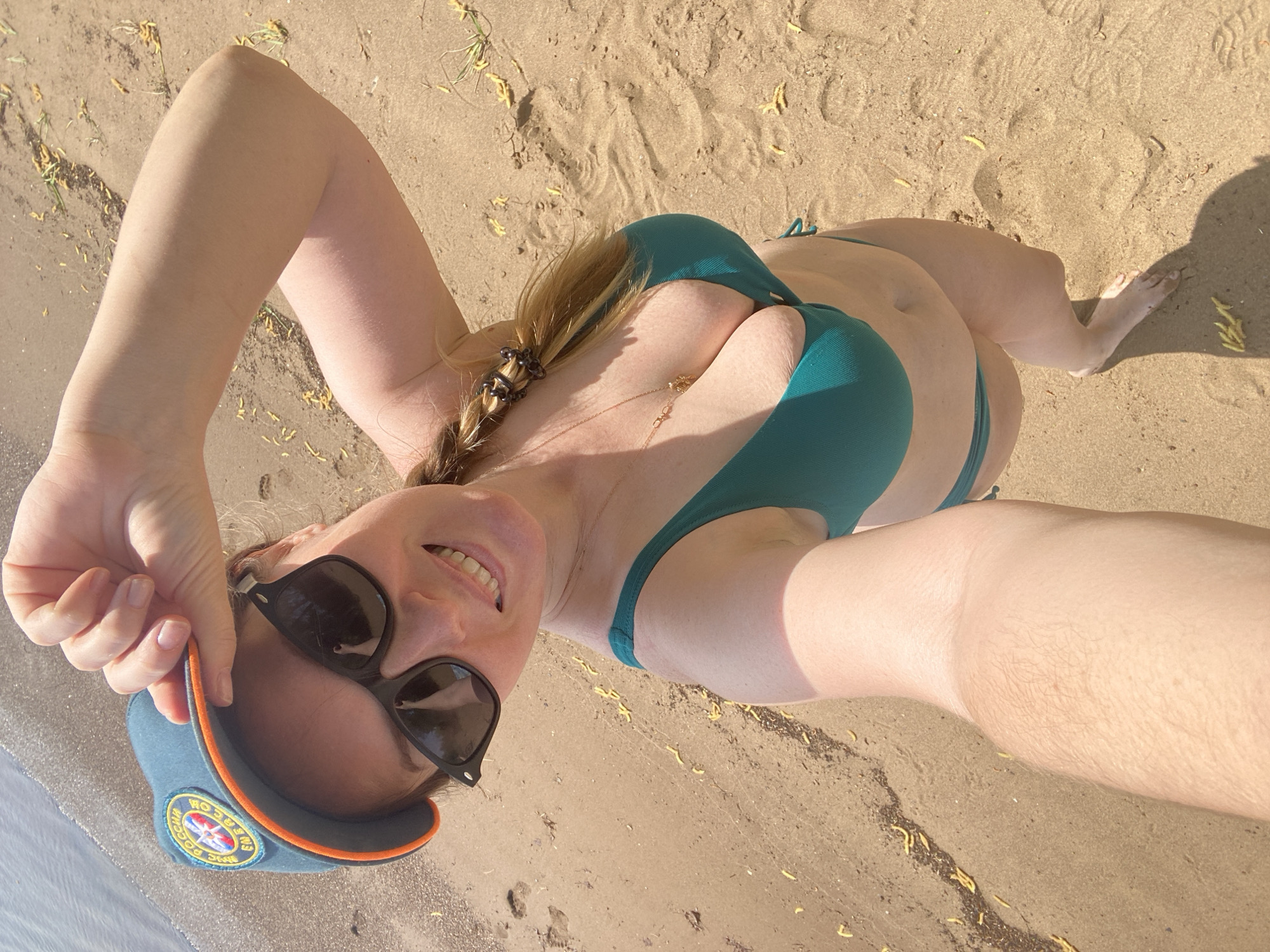 A woman is lying on a sandy beach, possibly near water. She is wearing a green bikini and sunglasses and is holding a cap with a logo on it. Her hair is braided. The location is identified as the city of Chaikovsky, Russia. The environment appears to be a beach or shoreline with light-colored sand.