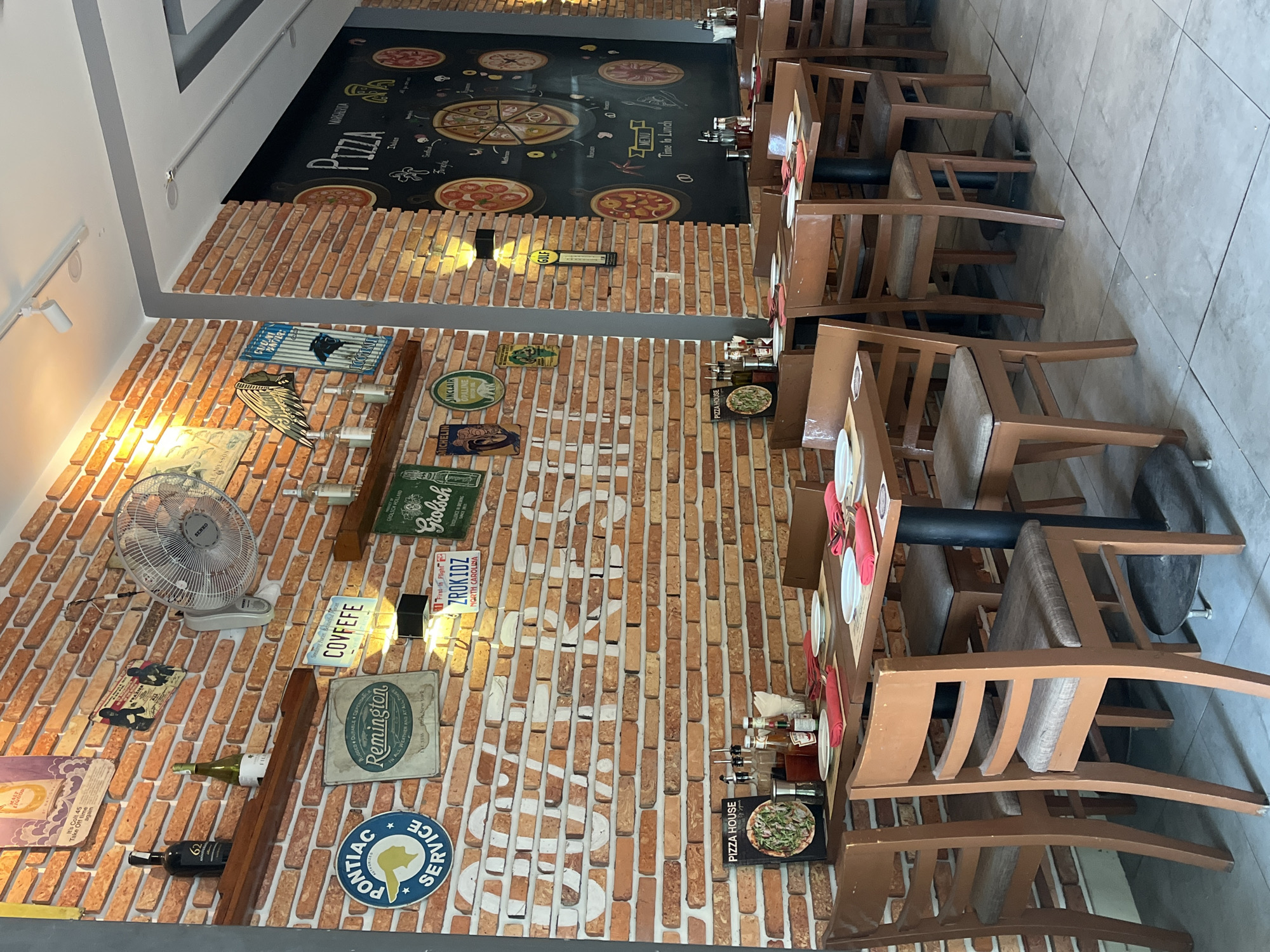 The image depicts the interior of a pizza restaurant in Đà Nẵng, Vietnam, furnished with multiple vacant dining tables and chairs. The walls are covered in a textured brick pattern, featuring white painted letters that spell "DA NANG" across a large section. Various vintage-style signs and decorations adorn the walls, including a circular "Pontiac Service" emblem, a rectangular "Remington" sign, a "Covfefe" license plate, a "ZROKIDZ NORTH CAROLINA" license plate, and signs for "Grolsch," "Michelin," "Sinclair Opaline," and "Gulf." Two wooden shelves are mounted on the left wall, holding various bottles, including a dark liquor bottle labeled "62" and a light-colored wine bottle. A white KDK wall-mounted fan is visible on the upper left wall.
On the right, a large blackboard display, framed by a brick wall section, presents hand-drawn pizza illustrations and text, including "PIZZA" at the top, and various pizza types like "MARGHERITA," "INDIAN," "SAUSAGE," "MUSHROOM," "ONION," and "MENU Time to Lunch." Several round pizzas are depicted with different toppings. The ceiling features track lighting and recessed spotlights.
The dining area consists of multiple wooden rectangular tables with dark metal pedestal bases, surrounded by wooden chairs with light brown upholstered seats. Each table is set with white plates, red cloth napkins, and cutlery. Condiment caddies containing bottles of ketchup, olive oil, and other sauces, along with salt and pepper shakers and napkin dispensers, are present on the tables. One table in the foreground displays a small "PIZZA HOUSE" menu board. The floor is covered in light-colored square tiles. No individuals are visible in the scene.
