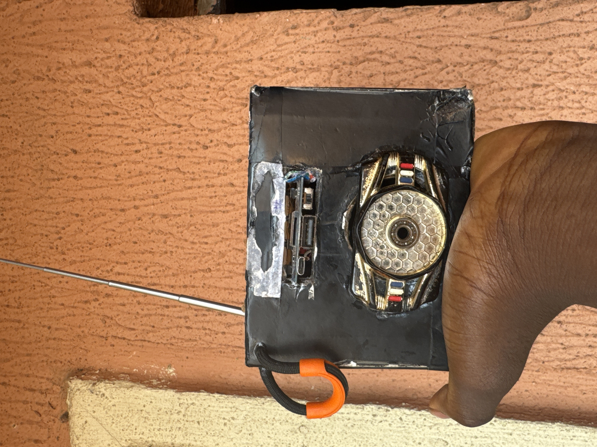 An overhead shot captures a dark-skinned hand holding a rectangular, likely homemade or repurposed electronic device against a textured, reddish-brown wall. The device is predominantly black, possibly covered in tape or plastic, and shows signs of wear with some peeling material.
The most prominent feature on the device is a circular, metallic component with a distinctive hexagonal pattern, resembling a lens or speaker grille, located on the right side. This component has gold-colored accents and what appear to be small red, white, and blue striped segments. To its left, exposed circuitry or internal components are visible through a torn or open section of the black covering. A thin, silver, telescopic antenna extends from the left side of the device towards the upper left corner of the frame. A black cord with a bright orange U-shaped clip or handle is attached to the bottom edge of the device.
The hand, with visible knuckles and skin texture, wraps around the right side of the device, suggesting it is being held for inspection, use, or display. The background is a rough, stucco-like wall, consistent with outdoor structures in places like Onitsha, Nigeria. Below the device and the hand, a section of a lighter, beige-colored wall or ledge is visible. The lighting suggests it is daytime. No discernible text is visible on the device or in the background. The scene depicts a casual interaction with a functional, possibly improvised, electronic item.