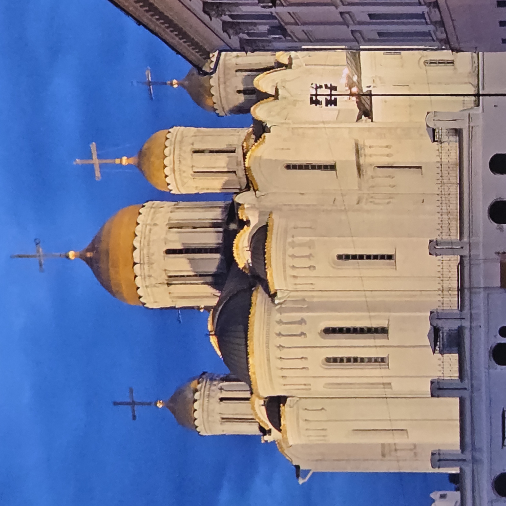 The image shows the Assumption Cathedral in Vladimir, Russia, against a deep blue twilight sky. The white stone cathedral features multiple golden domes, each topped with a cross. The architecture is characterized by white walls, arched windows, and decorative elements along the rooflines. A section of a modern building, appearing lighter in color with rows of windows, is visible to the right of the cathedral. The foreground shows a lower, lighter colored structure with an ornate fence and what appear to be arched openings at ground level. The scene is illuminated, suggesting artificial lighting is active.