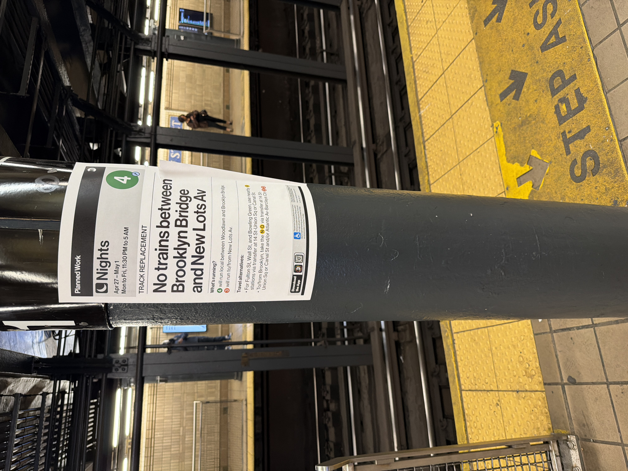 This image shows a sign posted in a New York City subway station. The sign, attached to a pole, informs passengers that there will be no 4 trains running between Brooklyn Bridge and New Lots Avenue due to track replacement. The notice specifies that this service change is in effect on weeknights from April 27 to May 1, between 11:30 PM and 5 AM.
The sign also details what train services will be running during the disruption, as well as travel alternatives for affected commuters. A yellow safety strip marked "STEP AS" is visible on the platform edge to the right. The station appears to be elevated, with train tracks and platforms seen through the station structure. In the background, two figures appear to be walking on a distant platform.