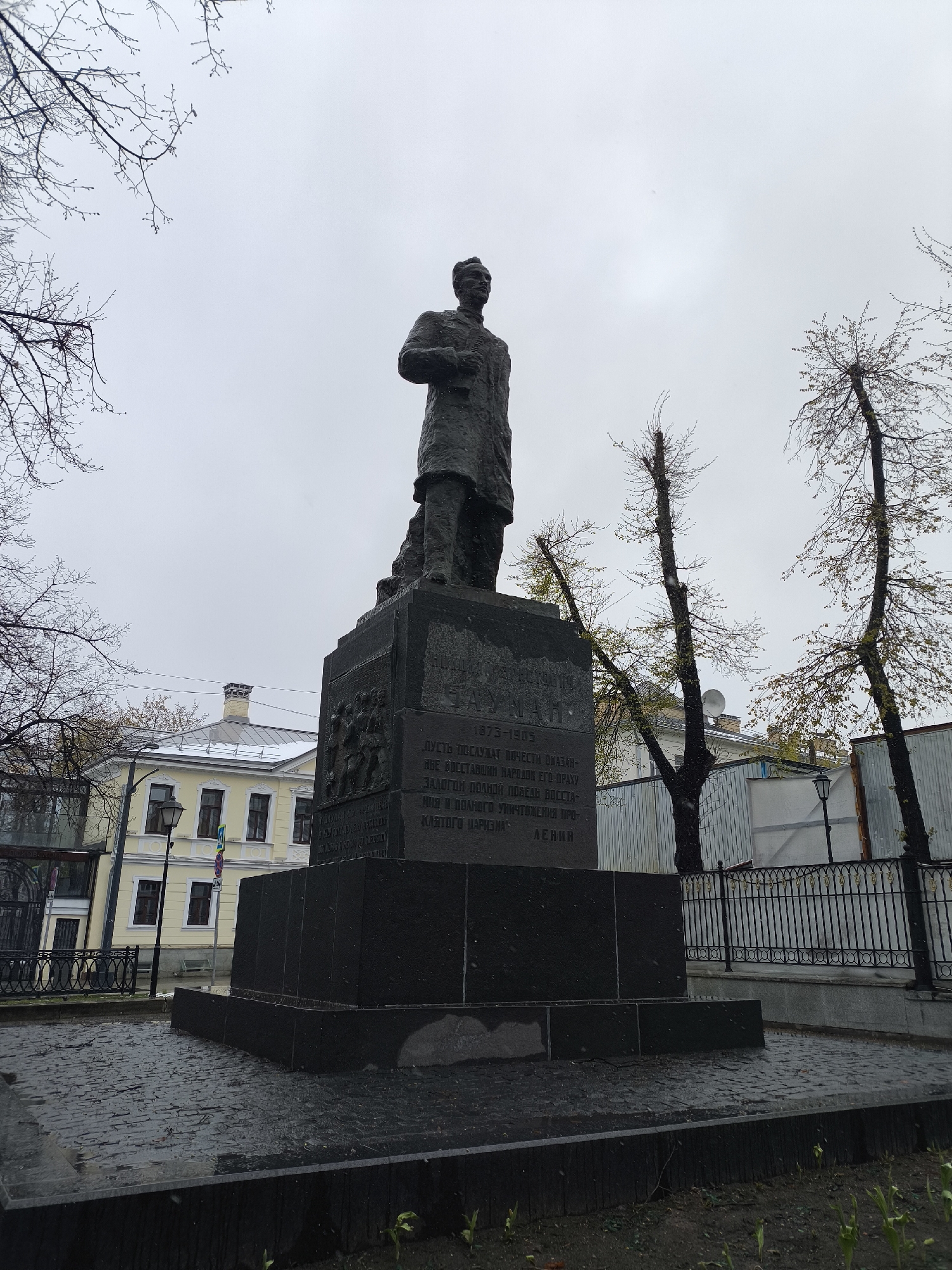 A bronze statue, likely depicting Vladimir Lenin, stands prominently on a dark, multi-tiered granite pedestal in an outdoor public space in Moscow, Russia. The figure is shown in a long coat, looking forward, with one hand held slightly forward. The base of the pedestal features bas-reliefs of figures, depicting historical scenes, possibly related to the revolution.
The scene takes place on an overcast day, with a muted, grey sky. The cobblestone ground surrounding the monument appears wet, suggesting recent rain or drizzle. Trees with bare branches or just beginning to sprout small, light-green leaves frame the monument, indicating early spring or late autumn. In the background to the left, a light-yellow building with dark windows and a snow-dusted roof is visible, characteristic of older Russian architecture. To the right, tall, pruned trees stand alongside a metal fence and more distant buildings. Two street lamps are also visible, one near the yellow building and another behind the fence on the right.
No specific activity or event is taking place; the image captures a static view of the monument.
The pedestal features Cyrillic text and dates. The visible text reads:
"1873-1905
ПУСТЬ ПОСЛУЖАТ ПОЧЕСТИ ОКАЗАН-
НЫЕ ВОССТАВШИМ НАРОДОМ ЕГО ПРАХУ
ЗАЛОГОМ ПОЛНОЙ ПОБЕДЫ ВОССТА-
НИЯ И ПОЛНОГО УНИЧТОЖЕНИЯ ПРО-
КЛЯТОГО ЦАРИЗМА.
ЛЕНИН"
This translates to: "1873-1905. Let the honors shown by the insurgent people to his ashes serve as a pledge of the complete victory of the uprising and the complete destruction of the cursed tsarism. LENIN." The quote is attributed to Lenin, reinforcing the identity of the statue. The dates "1873-1905" are present on the pedestal, possibly marking a period of revolutionary activity or another person's life, as they do not correspond to Lenin's birth and death dates (1870-1924).