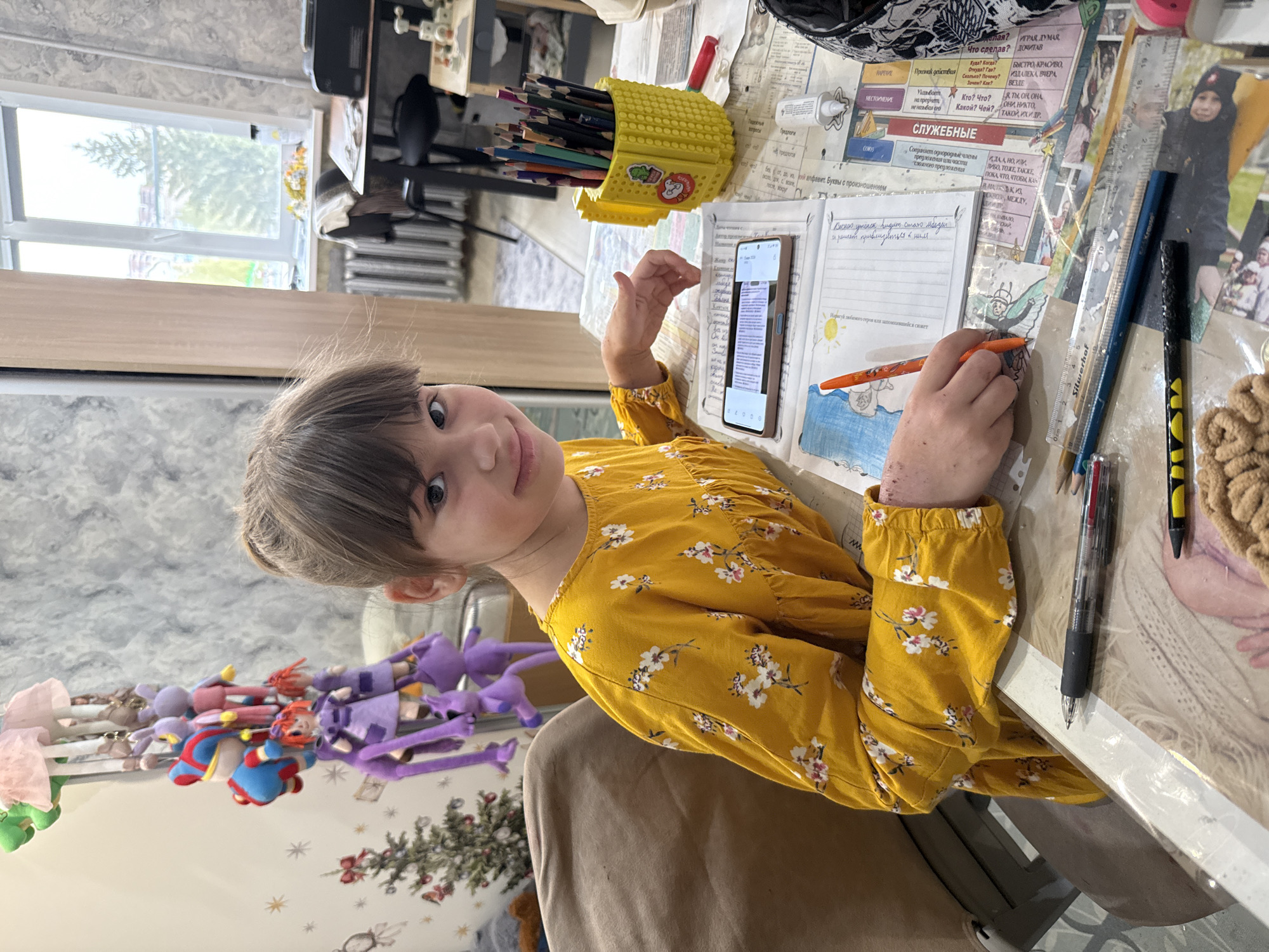 A girl is sitting at a table, looking at the camera. She is wearing a yellow dress with white flowers. On the table, there is a notebook, a pen, a phone, a pencil case filled with colored pencils, a ruler, a pencil, and some papers with writing in Russian. The notebook has a drawing of a winged figure. A picture can be seen taped on top of an article with words in Russian. A window and toys on a wall are visible in the background. The picture gives no indication of the location in the city of Rusanovo, Russia.