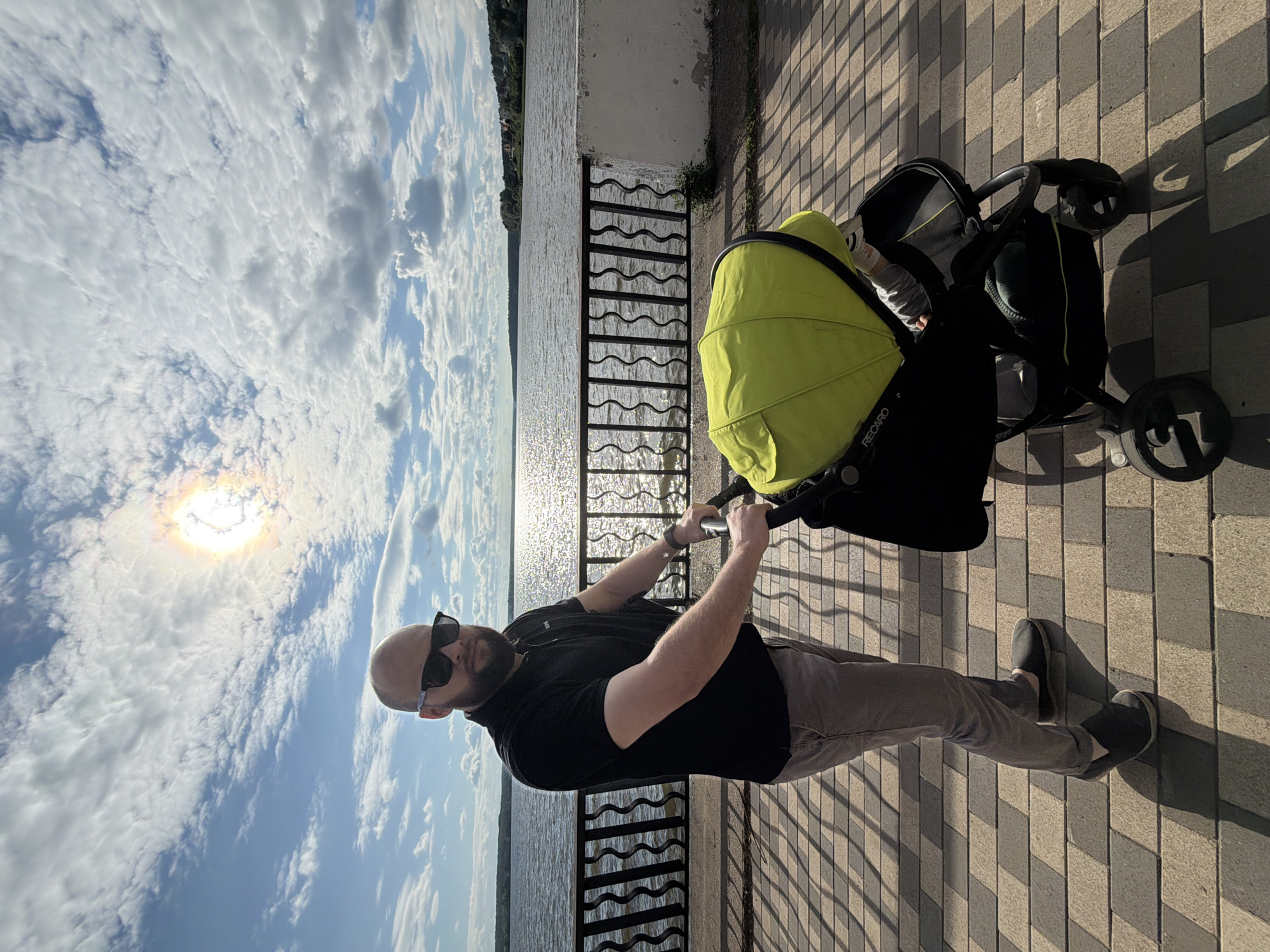 A man in sunglasses and a black polo shirt pushes a lime green stroller along a paved walkway. The walkway has a patterned brick surface. A dark metal railing with a wavy design separates the walkway from a body of water, likely the Votkinsk Reservoir in Votkinsk, Russia. The sun is visible in the sky, creating a lens flare effect and casting shadows from the railing onto the walkway. The sky is partly cloudy. In the distance, a shoreline with trees and some buildings is visible.