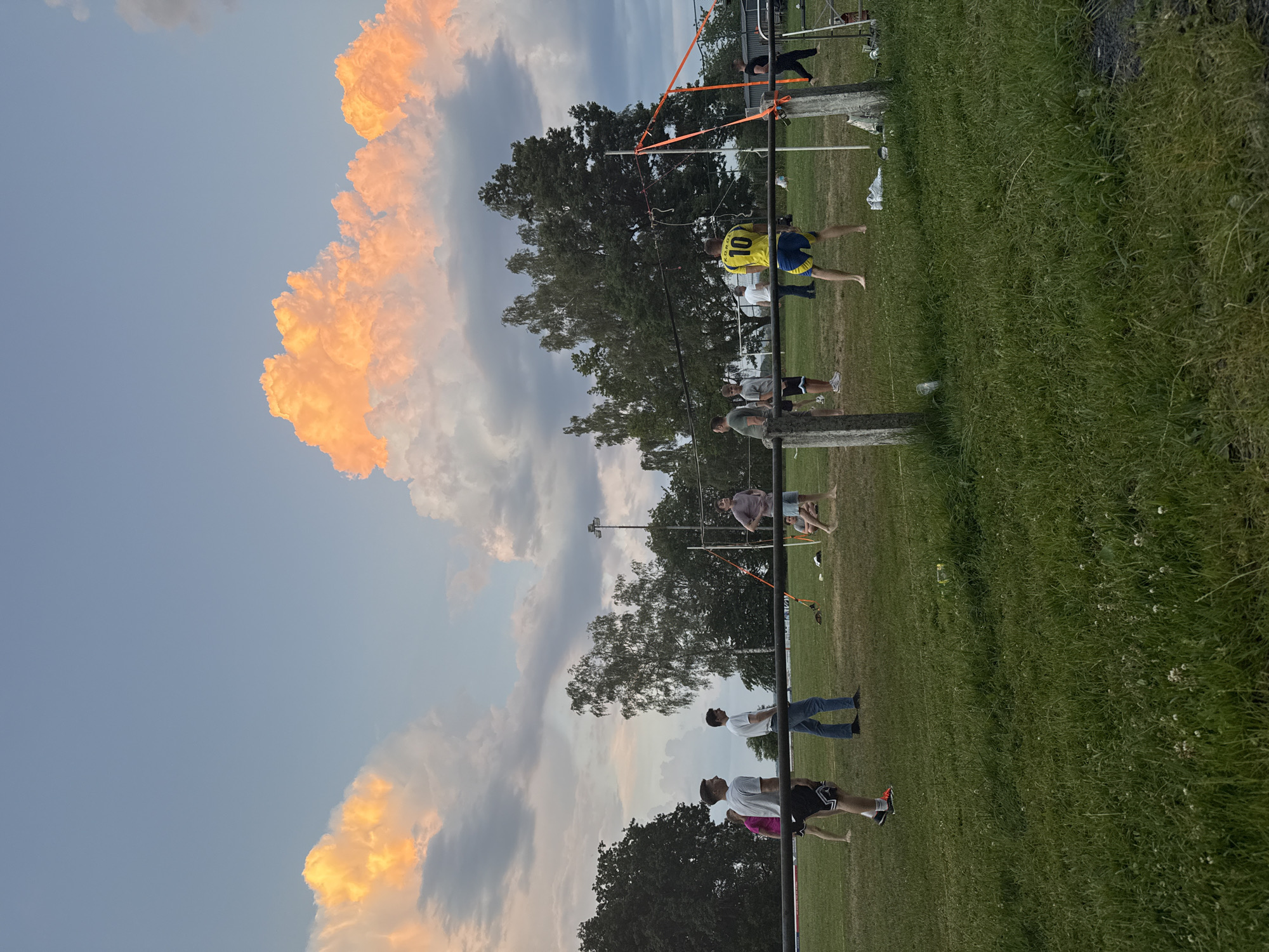 In Lumda, Germany, a group of young men are playing volleyball on a grassy field with a net set up across the middle. The sky above is dramatic, with large, fluffy clouds illuminated by the setting sun in vibrant shades of orange and pink. The overall atmosphere suggests a casual, outdoor recreational activity during the golden hour.