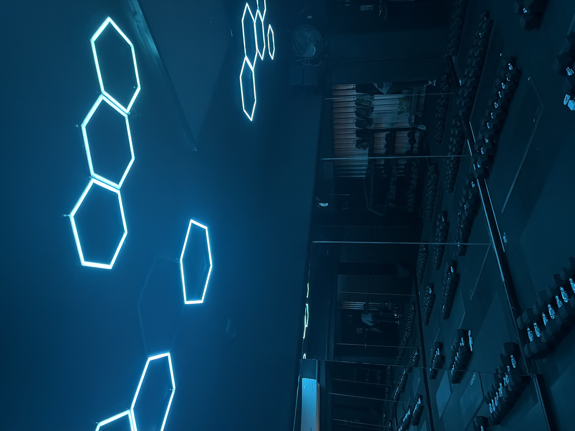 The image depicts an indoor scene, likely a modern gym or fitness center, characterized by its dark, blue-toned aesthetic and prominent neon lighting. The primary subjects are glowing cyan hexagonal lights arranged on a dark wall on the left side of the frame. These lights are clustered in various formations, with some extending out of view.
On the right side of the image, rows of gym equipment, specifically numerous dumbbells of varying sizes, are neatly arranged on racks. This area is heavily reflected, suggesting the presence of large mirrored walls, a common feature in gyms, which also reflect the hexagonal lights and other elements of the room. A ceiling fan is partially visible in the upper right quadrant of the image.
The overall color palette is dominated by deep blues and blacks, accentuated by the bright, luminous cyan of the hexagonal lights, creating a cool and potentially futuristic atmosphere. There is no visible human activity, but the setting clearly indicates a fitness environment. The time of day appears to be evening or night, given the reliance on artificial lighting.
A notable detail is the reflection quality, which provides a sense of depth and expands the perceived space. Visible text, though small and somewhat indistinct, appears to be "evelo" on a piece of equipment or label within the reflected gym area.