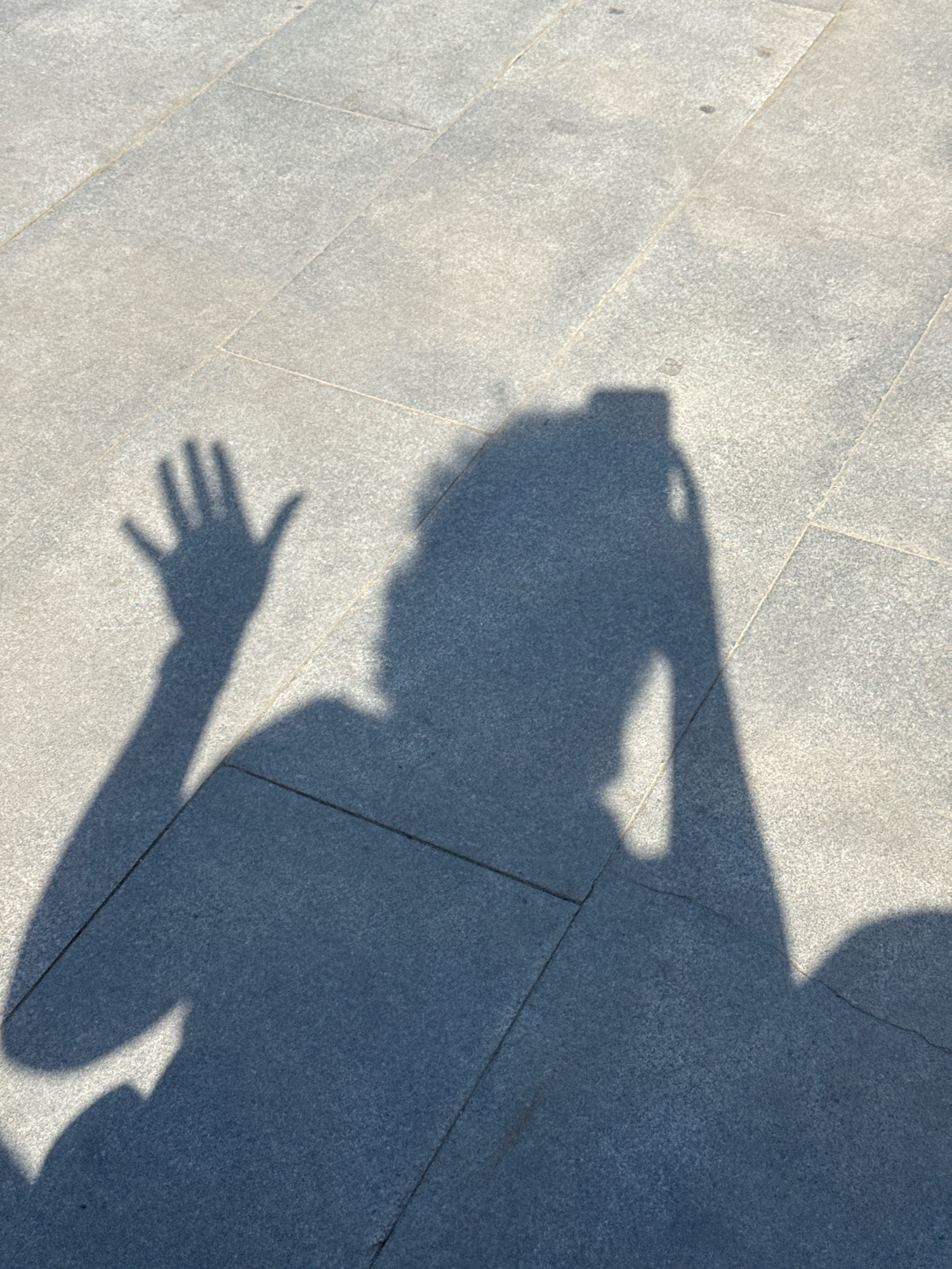 A shadow of a person taking a photo with their phone is cast on a paved outdoor surface in Yekaterinburg, Russia. The shadow depicts the person's head, torso, and an outstretched arm with a hand waving. The paving stones are visible, with distinct grout lines separating them. Sunlight is casting the shadow, indicating an outdoor daytime scene.