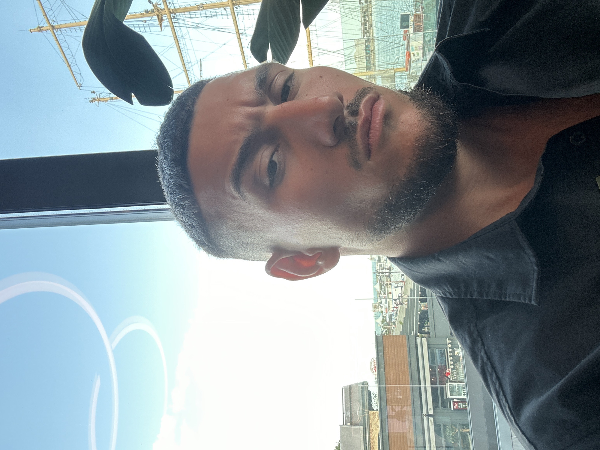 A man with dark hair and a beard is posing for a selfie indoors with a window behind him. The reflection in the window shows a tall ship docked in what appears to be a harbor. The visible landmarks suggest the city is Varna, Bulgaria, with the ship likely docked at the Varna Naval Museum and Harbour. The window also reflects buildings and signs along the waterfront. The man is wearing a dark-colored shirt. There's also a glimpse of a green plant near the top of the window.