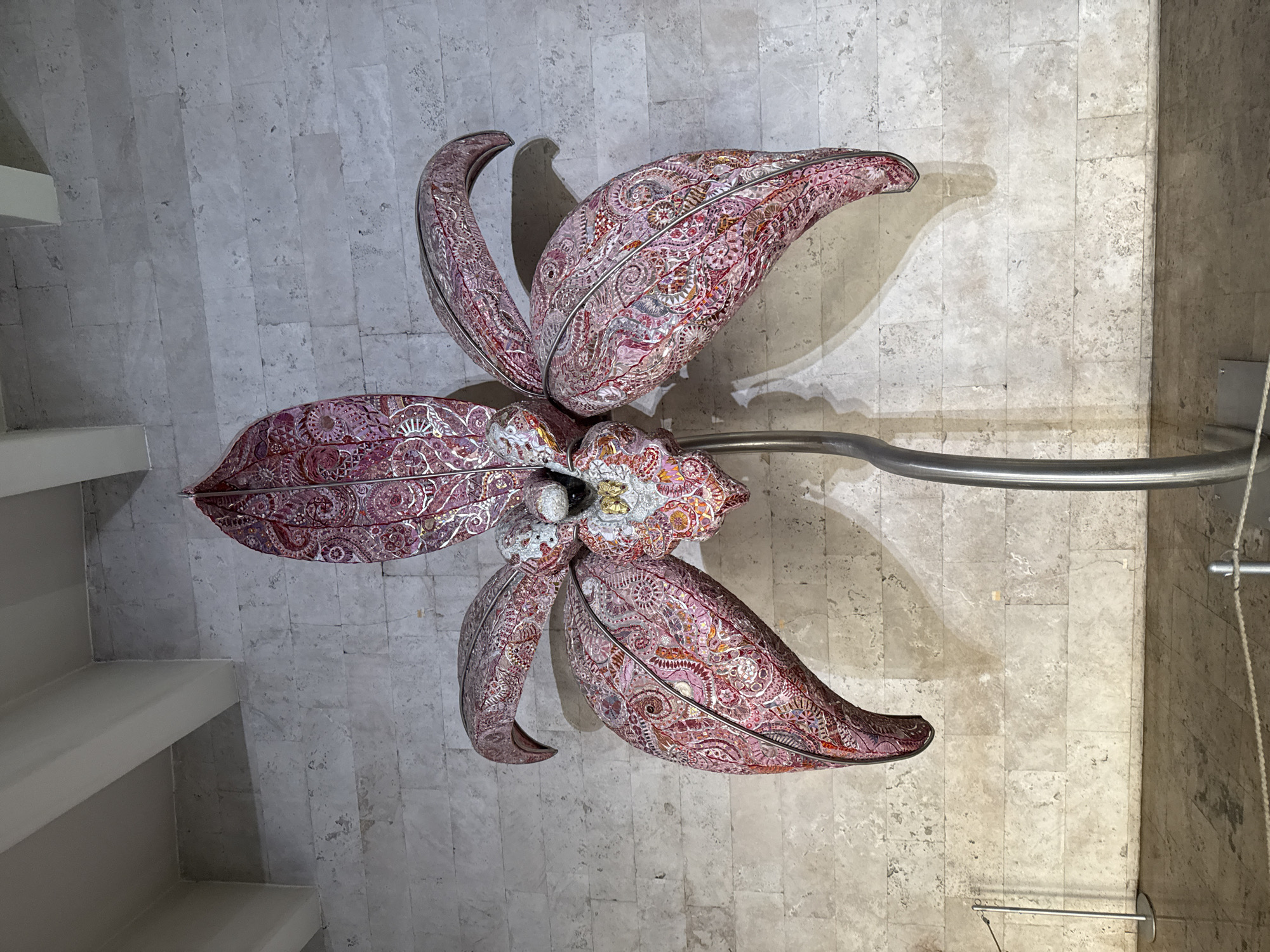 The image shows a large, ornate sculpture of a pink flower, possibly an orchid, displayed indoors against a textured, light-colored stone wall. The flower's petals are intricately decorated with detailed patterns and what appear to be small, reflective elements, giving it a mosaic-like appearance. The sculpture is mounted on a slender, curved silver stem, which is supported by a visible base. The lighting in the scene casts shadows of the flower and its stem onto the wall. In the foreground, a rope barrier is visible, suggesting a public space such as a museum or gallery. The floor is a smooth, reflective surface. The architecture of the ceiling, with its visible beams, suggests a modern or contemporary building. The sculpture is centrally framed and dominates the composition. This artwork is located in the Cafesjian Center for the Arts in Yerevan, Armenia.