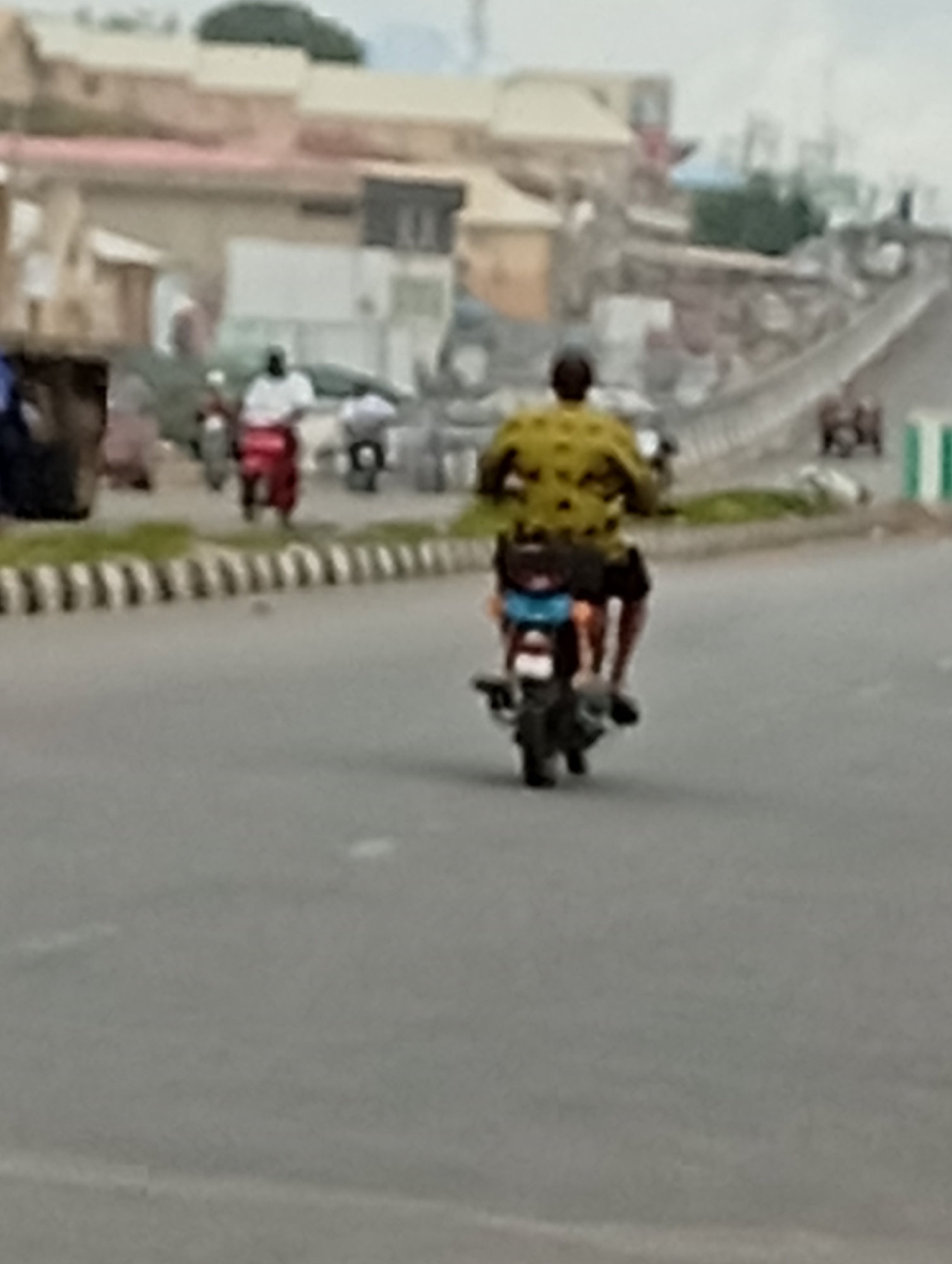 A person rides a motorcycle down a street in an urban environment. The rider wears a yellow shirt with a dark pattern. The motorcycle has a blue object on the back. In the background, there are buildings and an overpass. Other motorcycles and people are visible further down the street. The street appears to be paved, with a striped barrier along the side. The sky is overcast.