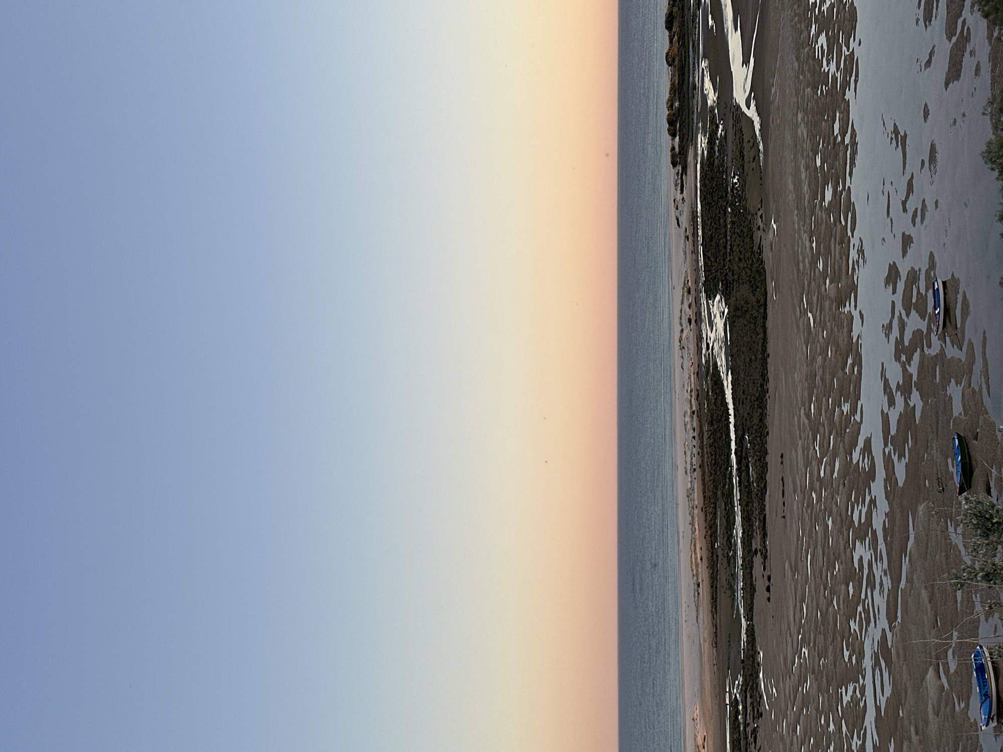 The image depicts a coastal scene at low tide. A wide expanse of wet sand is visible, reflecting the sky and creating mottled patterns. Two small boats are beached on the sand. A strip of vegetation runs along the edge of the sand. In the distance, the sea meets the sky, with a gradient of colors from blue to orange. The location is likely in Vila Real de Santo António, Portugal, as suggested by the prompt, given the coastal geography and tidal range.