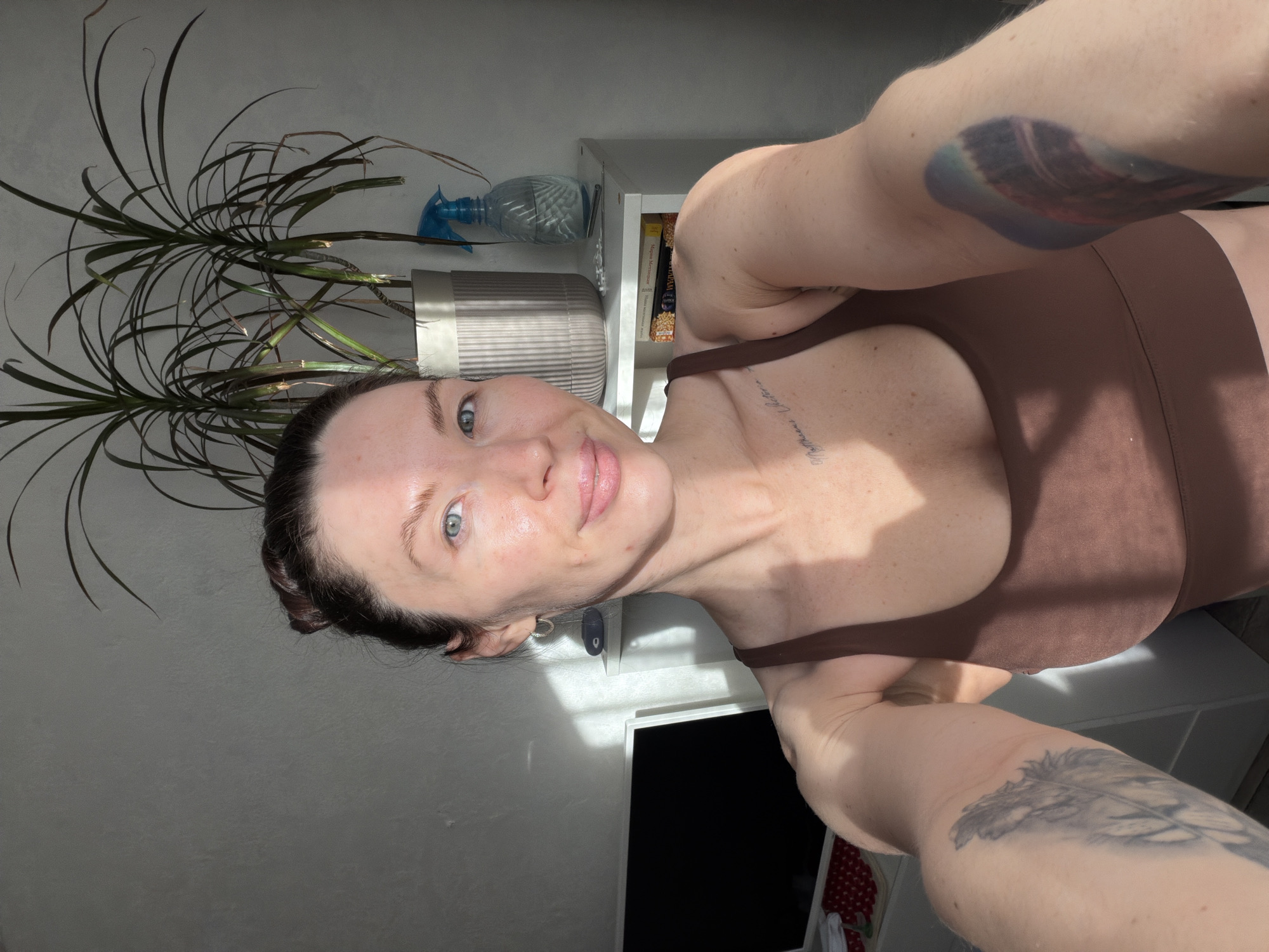 A fair-skinned woman with blue eyes and dark hair is taking a selfie. She is wearing a brown top and has tattoos on both arms and her upper chest. In the background, there's a potted plant, a white shelf with a spray bottle, and some books. The room is brightly lit, possibly by sunlight. There is no information in the image to determine if this image was taken in Perm, Russia.