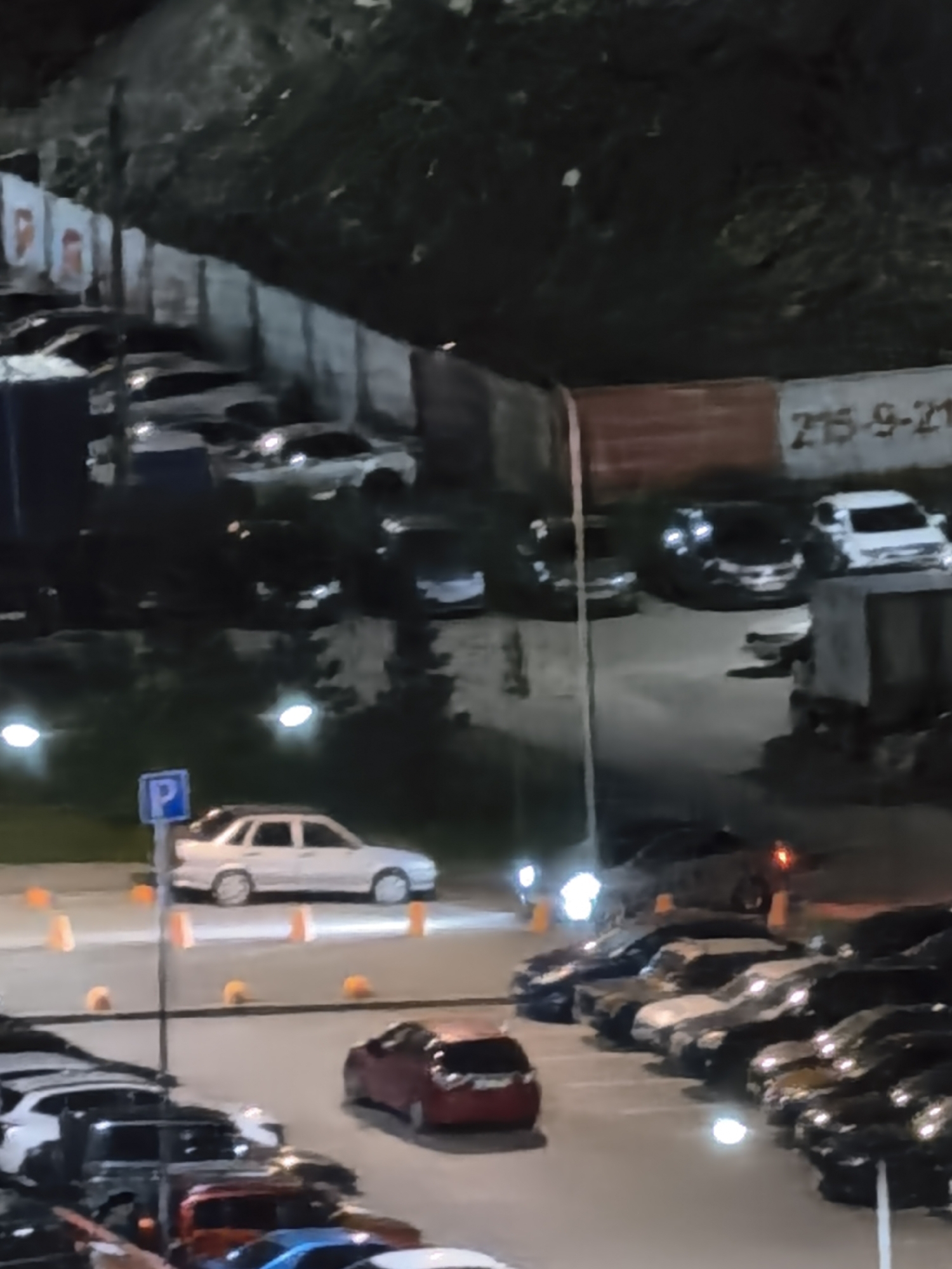 The media file shows a nighttime view of a parking lot filled with cars. The scene appears to be outdoors, possibly in an urban or suburban area, given the presence of a streetlamp in the background and markings on the pavement. The lighting is dim, with the headlights of some cars illuminating the area, and there are some orange traffic cones scattered around.
The main subjects are the parked cars of various colors and models. There are no people clearly visible in the image. The overall activity is simply the presence of a parked car collection.
Notable details include the time of day being nighttime, indicated by the darkness and artificial lighting. The weather is not discernible. There are no strong emotional cues evident from the static image.
Visible text includes what appears to be a parking sign with a blue "P" on a white background. In the upper right portion of the image, there is a segment of text on a wall that reads "275-9-21". The overall setting suggests a typical parking area, perhaps after business hours or in a residential zone.