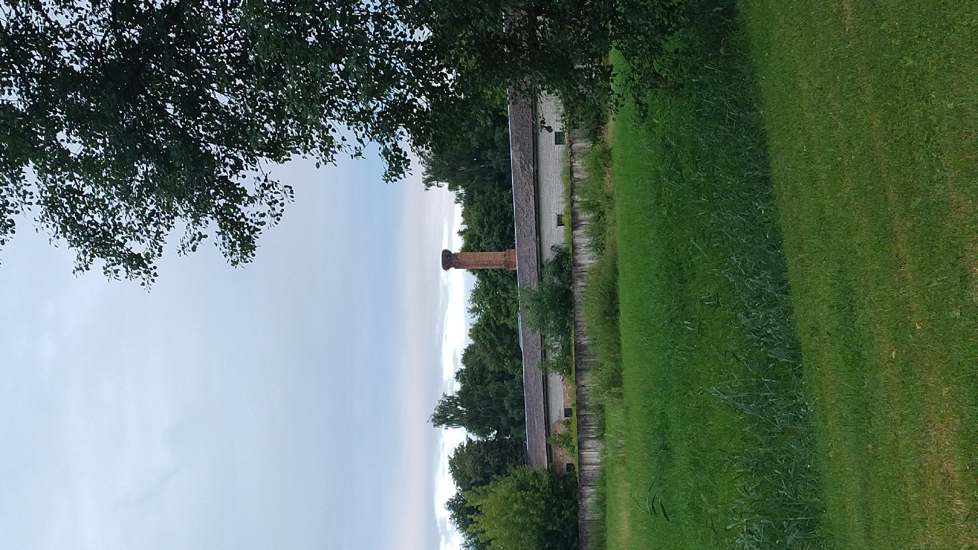 The image depicts a pastoral scene with a long, low building with a grey roof in the middle ground, partially obscured by trees. A tall, red brick chimney stands behind the building. A concrete fence runs along the front of the building, with tall green grass in the foreground. The sky is overcast. This scene is consistent with the landscape found on the outskirts of Jelgava, Latvia, where agricultural buildings and open fields are common.