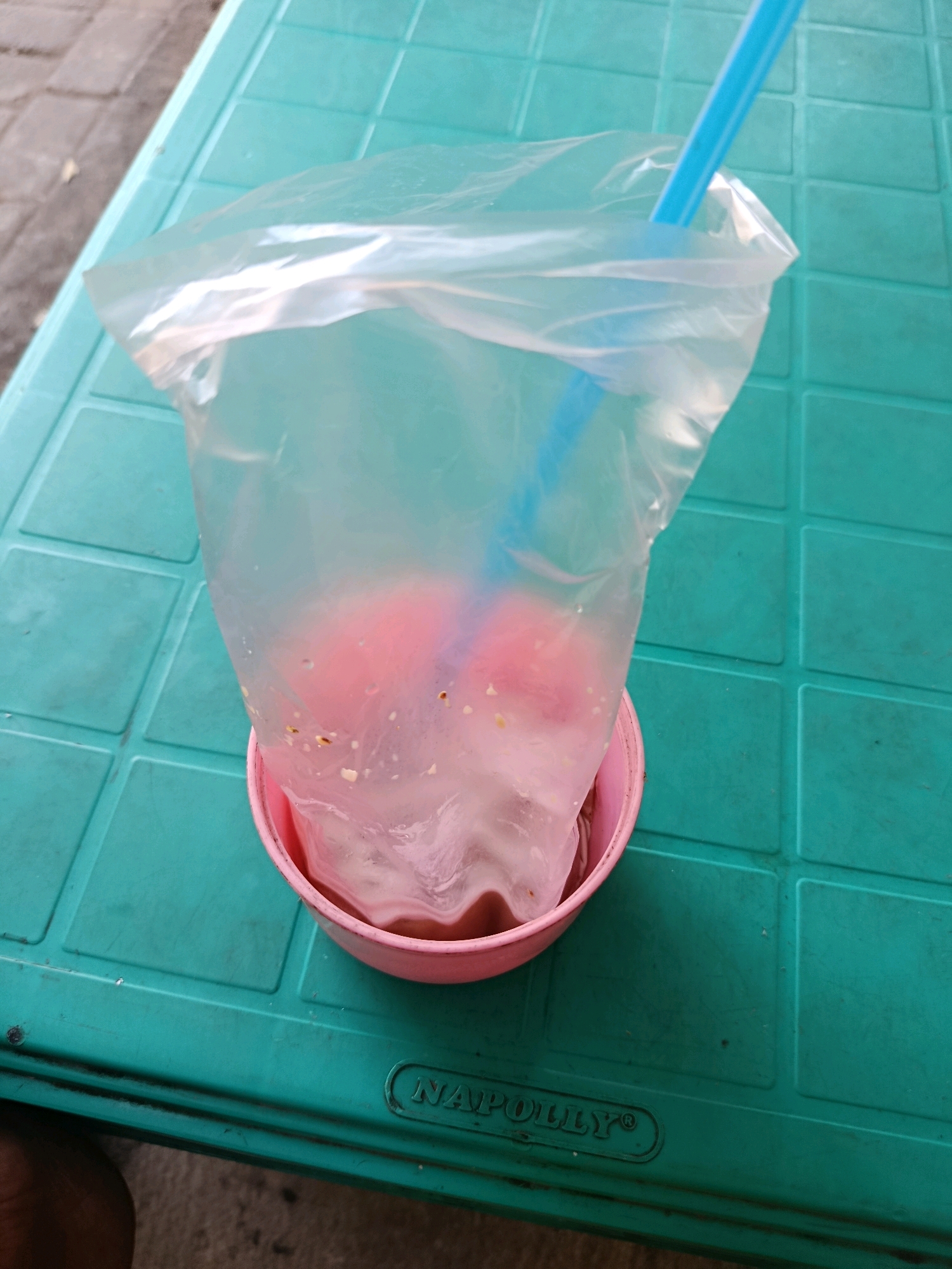 A plastic bag containing a pink beverage and ice is resting in a pink bowl on a teal-colored, grid-patterned table. A blue straw extends from the bag. The table has the embossed text "NAPOLLY" on its surface. The background shows a cobblestone-like surface, possibly a street or sidewalk. The scene appears to be outdoors, possibly in Jobokuto, Indonesia.
