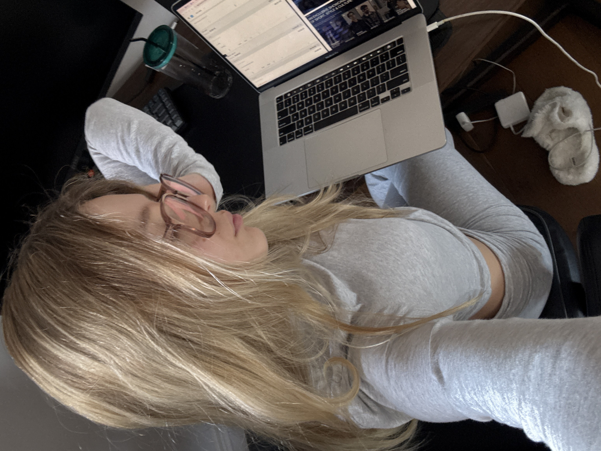 A woman with long blonde hair lies reclining in an office chair wearing glasses and a light gray, long-sleeved shirt and matching sweatpants. A laptop sits on her lap, displaying a screen with tabular data on the left and an image of people on the right. To the left of the laptop is a partially filled clear plastic cup with a green lid and straw. A black keyboard is also visible in the background. A power adapter, charging cable, and a pair of fuzzy slippers are visible on the floor to the right of the laptop. The location is unknown and cannot be determined from the image.