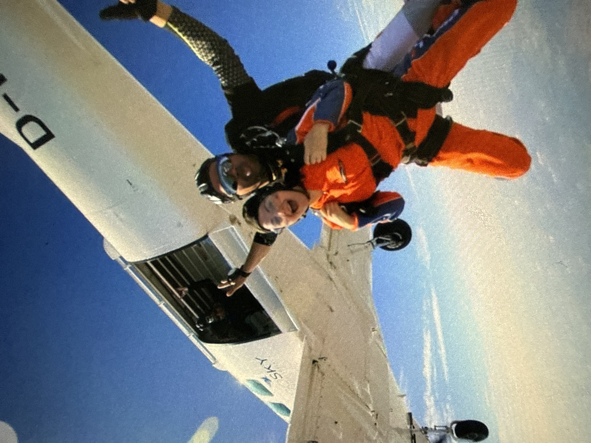 Two people are skydiving in tandem out of an aircraft. One person is wearing an orange jumpsuit and the other is wearing a black suit with speckled sleeves. The aircraft is white with the word "SKY" printed on it. The location is in the city of Unknown, Unknown. The sky is blue.