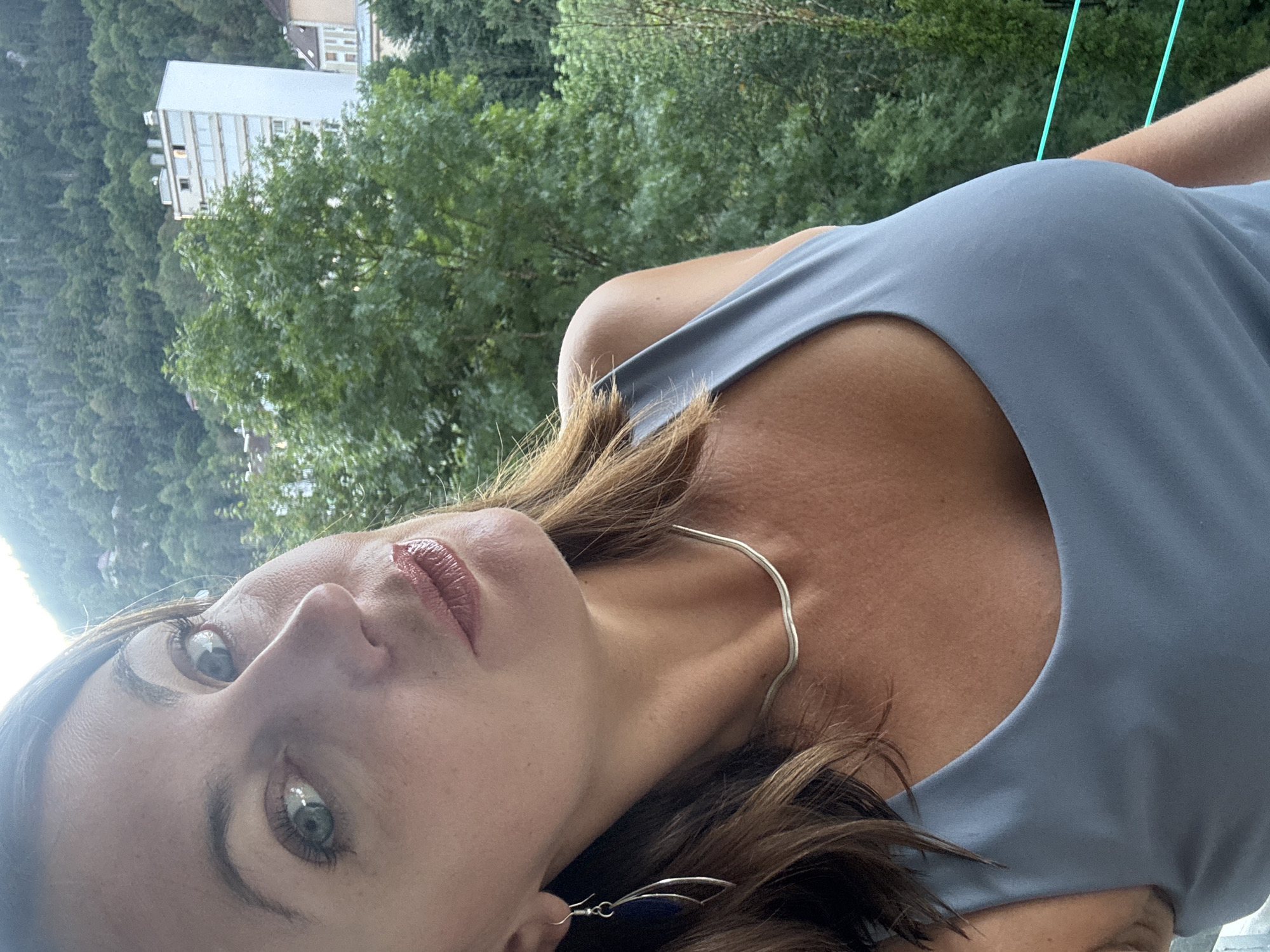 A woman is captured in a close-up, slightly angled selfie taken from the chest up, with a blurred background of lush green trees and a white building, indicative of the city of Saint-Claude, France. The woman has light skin, brown hair that falls around her shoulders, and is wearing a slate-colored tank top, a delicate silver necklace, and a long silver earring with a dark blue stone. Her head is tilted to the left, and she is looking towards the camera with her eyes open. Her lips are slightly parted and appear to be colored with a glossy lipstick. The background shows a dense forest climbing a mountainside, with a modern white building standing out amongst the trees. The overall impression is a casual portrait taken outdoors, likely on a balcony or elevated vantage point, offering a glimpse of the natural beauty and urban landscape of Saint-Claude.