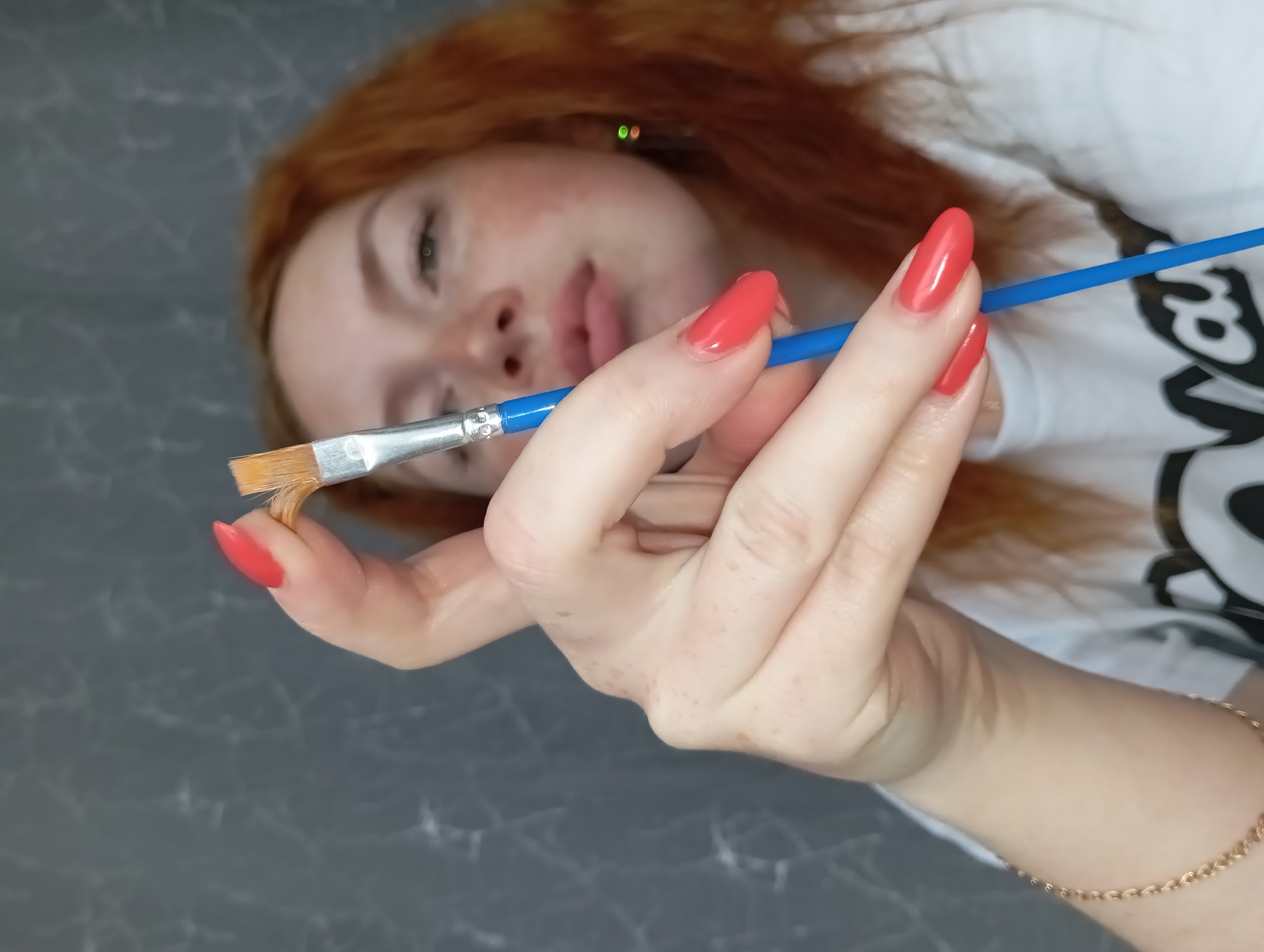 A pale-skinned, red-haired woman with freckles and red nail polish is holding a blue paint brush. She wears a white t-shirt with black print and a gold bracelet. She appears to be indoors against a grey background. There is no other information to indicate this image was taken in Yurgamysh, Russia.