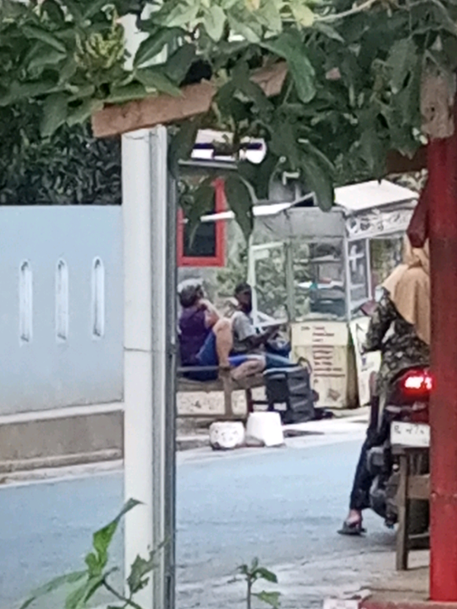 In Kalikidang, Indonesia, an outdoor scene unfolds with several individuals and elements of a street-level environment. In the foreground, lush green foliage partially obscures the view. To the left, a light blue wall with three vertical white rectangular indentations is visible. In the mid-ground, two individuals are seated on a bench or low structure. One person, wearing a purple top and blue shorts, is turned away from the camera, with their legs crossed. Next to them, another person, wearing a grey t-shirt and blue pants, is also seated and appears to be looking towards the right. Behind them, a small, open-air stall or kiosk with transparent walls displays various items on shelves. A black bag and a white bucket are situated on the ground near the seated individuals. Further to the right, a person wearing a camouflage patterned outfit and a tan headscarf is seated on a motorcycle, facing away from the camera. The motorcycle's brake light is illuminated. The background consists of a building with a red-framed window. The overall setting suggests a street or walkway with elements of commerce and daily life.