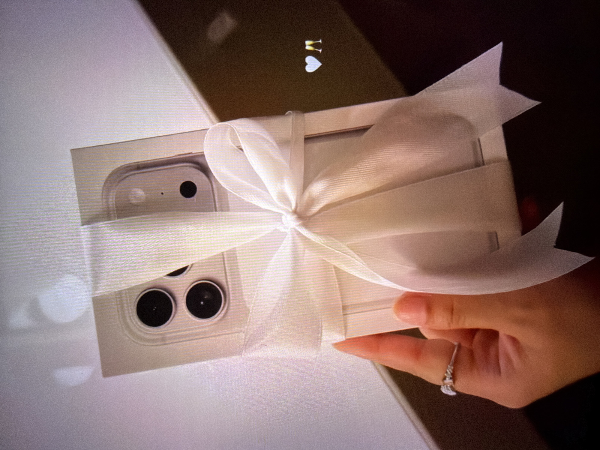 A close-up shot shows a hand holding a white gift-wrapped phone box. The box is adorned with a wide white ribbon tied in a bow. The phone's camera lenses are visible through the ribbon. A ring is on the index finger of the hand. In the background, there are two small icons: a white heart and two champagne flutes. The image appears to have been taken indoors, possibly in a home setting. There is no visible information indicating a specific location within Beșalma, Moldova.