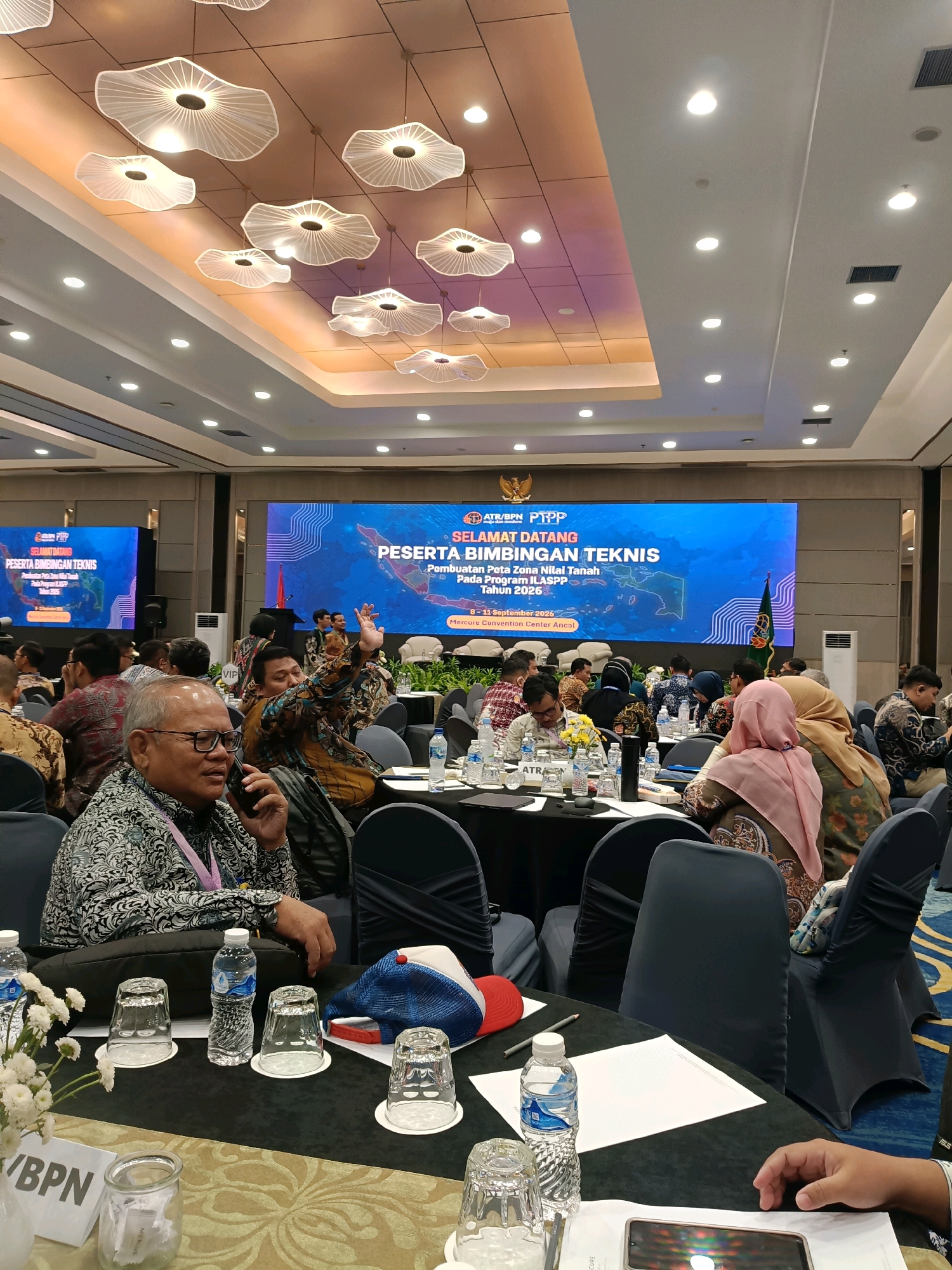 The image depicts an indoor conference room in North Jakarta, Indonesia, likely at the Mercure Convention Center Ancol, as suggested by a sign visible on the table in the foreground. The room is set up for a seminar or workshop, with round tables arranged for attendees. A large digital screen at the front displays "SELAMAT DATANG PESERTA BIMBINGAN TEKNIS PEMBUATAN PETA ZONA NILAI TANAH Pada Program ILASPP Tahun 2026".
In the foreground, a man wearing a patterned shirt and glasses is speaking on a mobile phone while seated at a table. On the table, there are water bottles, glasses, and a cap. Other attendees, some wearing traditional Indonesian attire, are seated at various tables around the room. The ceiling features modern, decorative lighting fixtures. The overall atmosphere suggests a professional event or gathering.