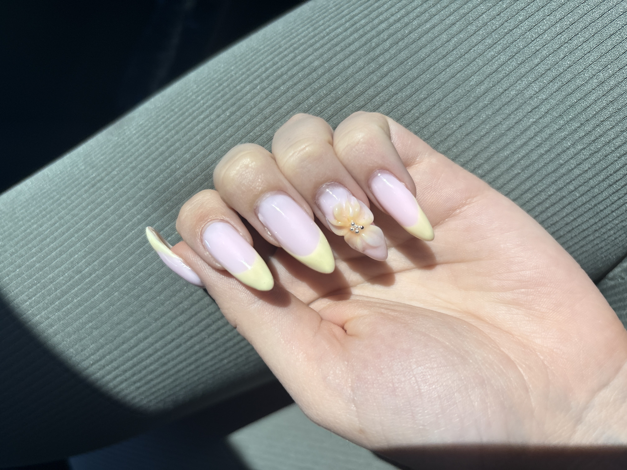 A close-up of a person's hand, with long nails painted in a French manicure style with pale pink base and pastel yellow tips, except for one nail decorated with a 3D flower and clear rhinestones. The hand is resting on a textured, gray surface, likely a car seat. The scene appears to be illuminated by natural light. The location in the city of Burgas, Bulgaria is not discernible from this image.