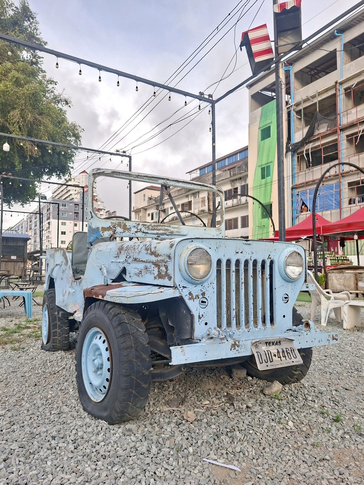 In Pattaya City, Thailand, an old, light blue Jeep Wrangler sits in a gravel lot. The Jeep is heavily weathered, with peeling paint and visible rust, suggesting it is no longer in operational condition. The vehicle's tires are flat, further indicating it's a static display. In the background, buildings of varying heights and architectural styles line the street, some appearing under construction. Red awnings are visible over what seems to be an outdoor market or seating area. Overhead, string lights are suspended, hinting at a casual outdoor venue. The sky is overcast.