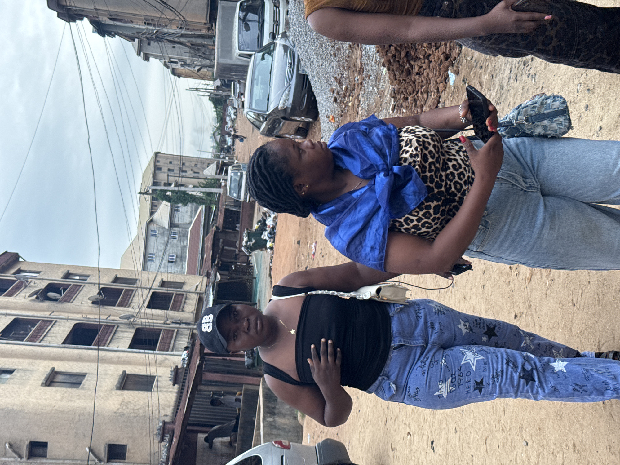 The image shows two Black women standing on a dirt street in Onitsha, Nigeria.
The woman on the left wears a black tank top, blue jeans with star patterns, and a black baseball cap. She has a gold necklace and a small white purse. She is holding her hand to her chest and looking at the camera.
The woman on the right wears a leopard print top, a blue scarf, and light blue jeans. She is looking at a phone in her hands. A small denim purse is visible next to her. Another woman is standing to the right of her and we can only see her lower body.
Behind the women, there are buildings, cars, and gravel on the side of the road. Overhead electrical wires are visible. The sky is overcast.