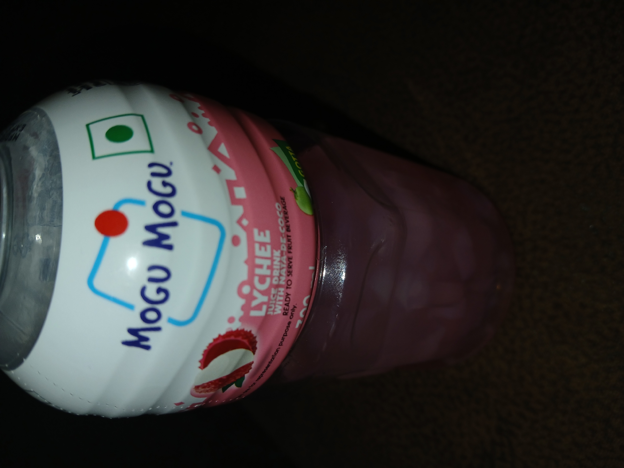 A single bottle of Mogu Mogu Lychee juice drink is viewed from above and slightly to the side. The bottle has a white and pink label, with the brand name "Mogu Mogu" prominently displayed. The label also features a lychee fruit graphic and the text "LYCHEE JUICE DRINK WITH NATA-DE-COCO READY TO SERVE FRUIT BEVERAGE". The bottle appears to be resting on a dark surface. The location is unknown, and there is no verifiable indication of being in Bengaluru, India.