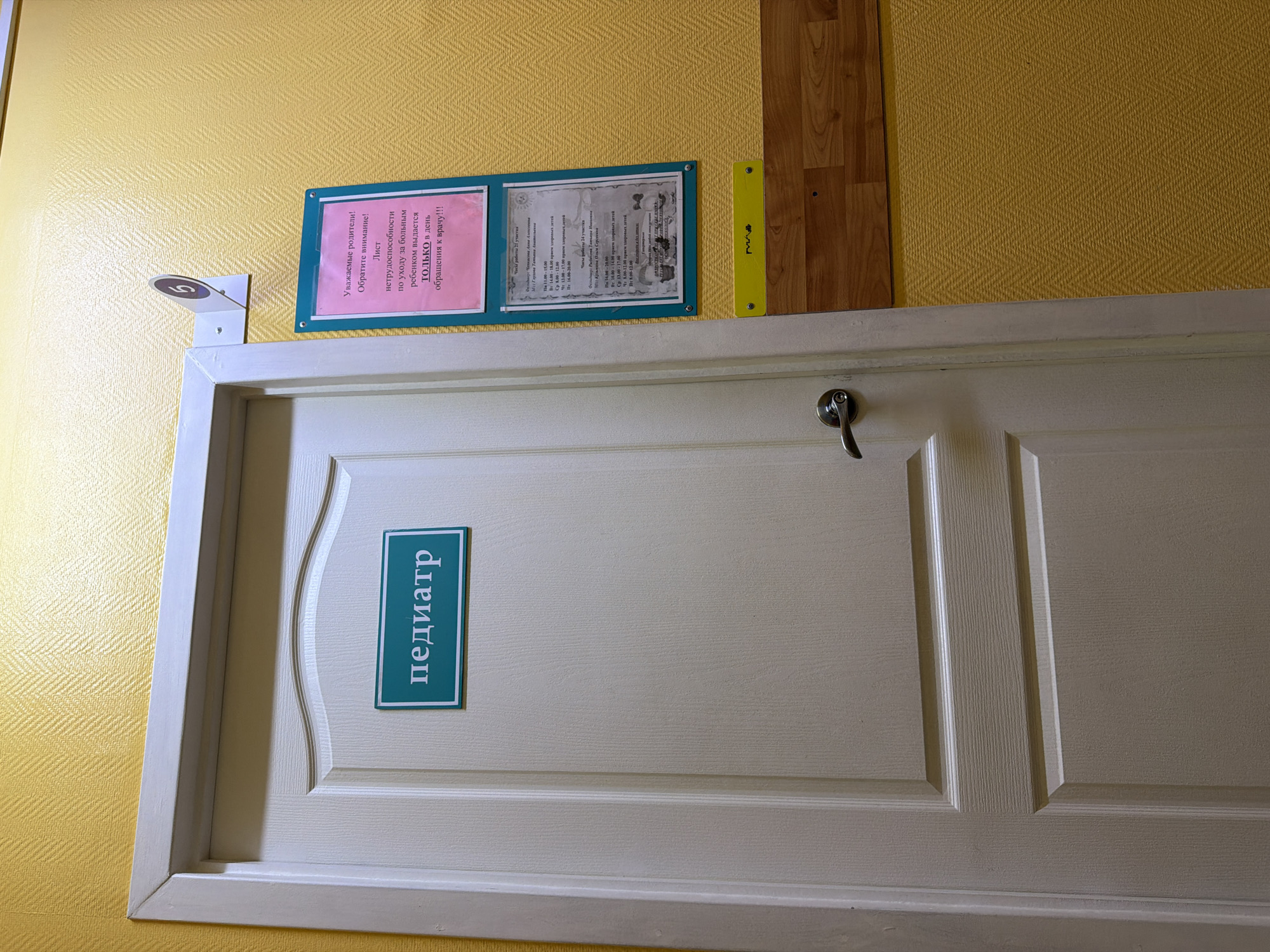 A mostly centered, medium shot depicts the top portion of a white door labeled "педиатр" (pediatrician) in teal vertical lettering, likely in Chaikovsky, Russia. A silver handle is on the door's right side. Above the door, on a yellow wall with a textured pattern, are two signs with Russian text mounted in a blue frame. The sign on the left is pink and cautions that disability certificates for childcare are issued only on the day of the visit. The sign on the right lists clinic hours for two area pediatricians. To the left of the signs is a white metal plate with a number 5 on it. To the right of the signs is a strip of faux wood paneling. The lighting is bright but somewhat uneven, with shadows suggesting an artificial light source.