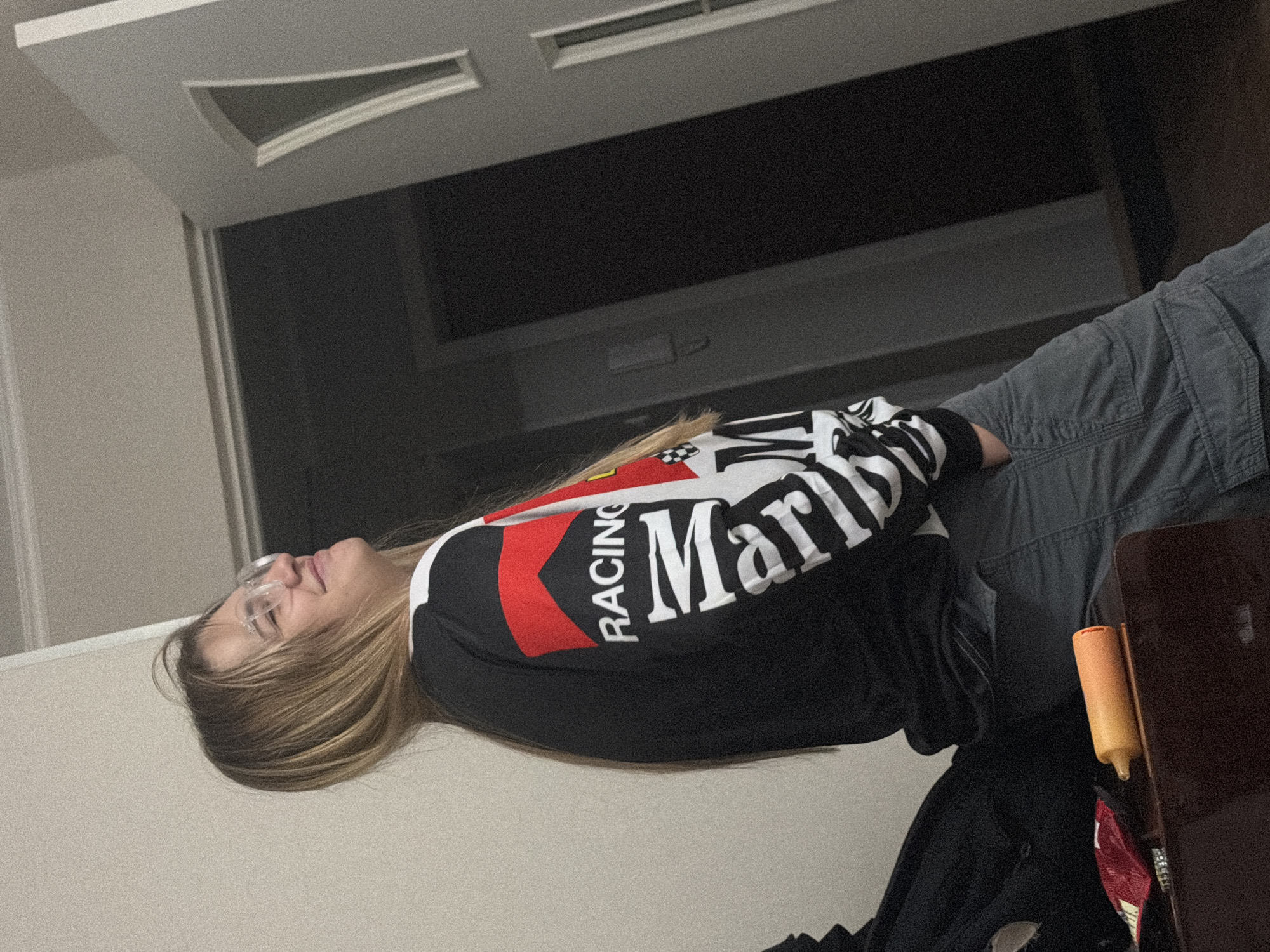 A medium shot shows a woman in a Marlboro racing jacket and grey pants. She wears glasses, and her hair is blonde with dark roots. The woman is lying on a light beige surface. A partially visible, dark wooden table with an orange object is present in the lower right corner. The background features a white door with a triangle design and a dark window. The image's location is unknown and no elements indicate the image was taken in Kazan, Russia.