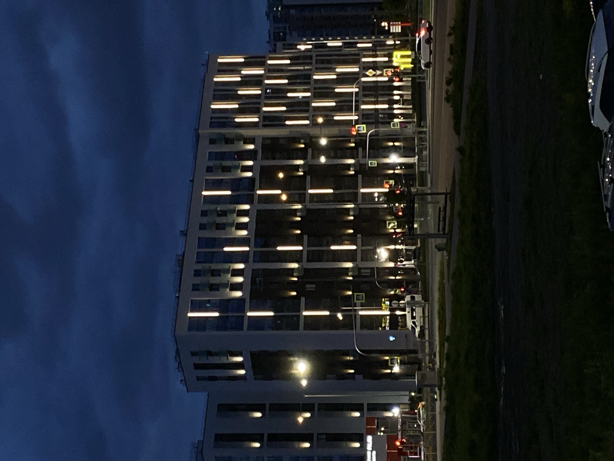 An exterior shot, taken at night, shows a tall building in Saint Petersburg, Russia. The building is illuminated with horizontal lines of light coming from many of its windows. Street lights also appear. To the right of the building is a road, and beyond that, a grassy area. Several vehicles, including cars and vans, are visible on the road. The sky is dark.