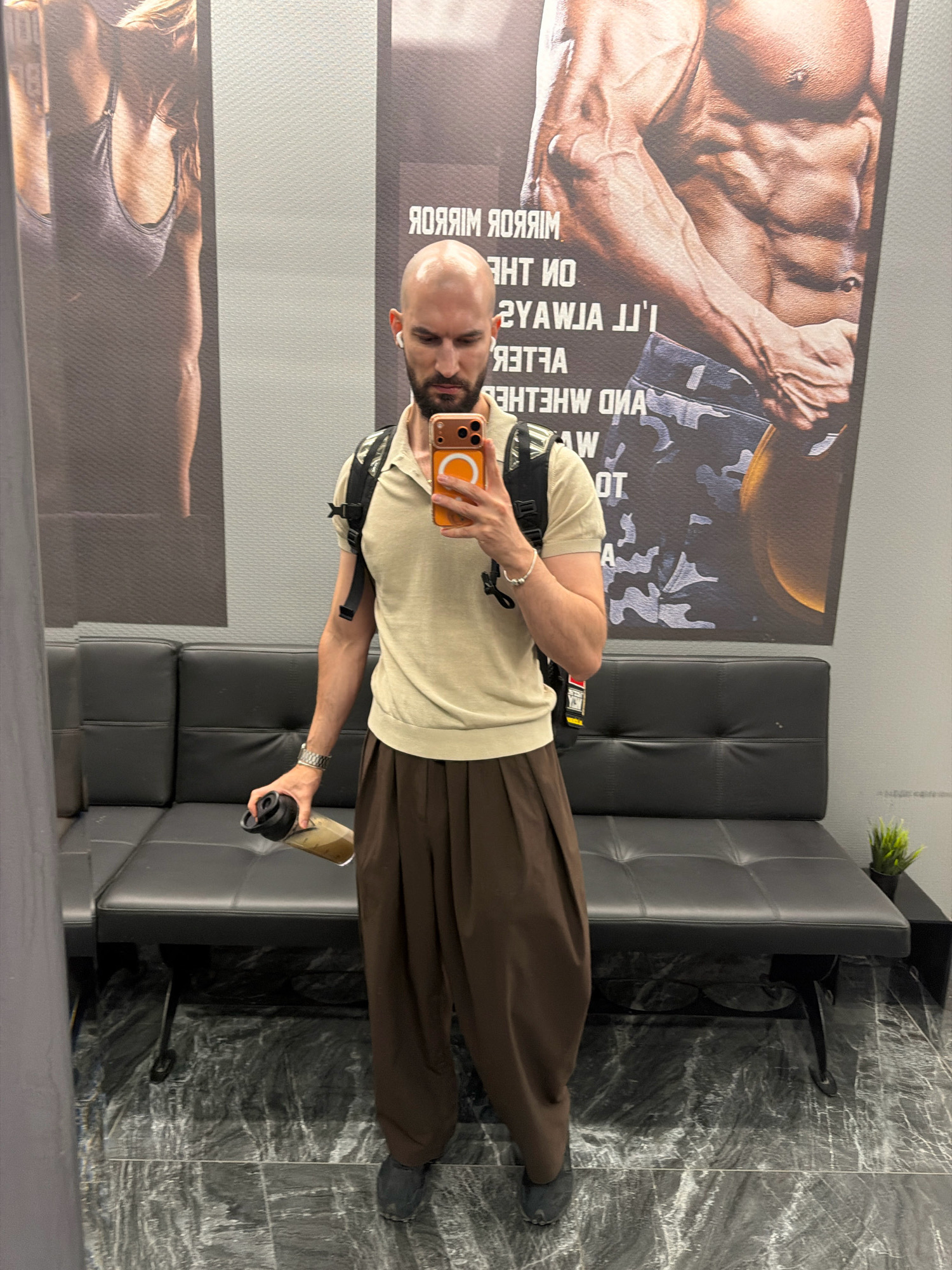 In a gym locker room, a bald, white man with a beard and white earbuds takes a mirror selfie. He wears a light beige polo shirt, loose brown trousers, and dark shoes. A black backpack with red accents is on his shoulders. In his right hand, he holds a black water bottle with a translucent lid, containing a greenish liquid. He is standing in front of a black leather bench, with a large motivational poster of a muscular man's torso on the wall behind him. The floor is tiled with a dark, marble-like pattern. The man's reflection is visible in a mirror to his left, showing part of his arm and torso. To the right, another poster features the back of a woman's muscular physique. A small green plant sits on a black stand next to the bench on the right. The environment suggests a public fitness facility, possibly in Sofia, Bulgaria.