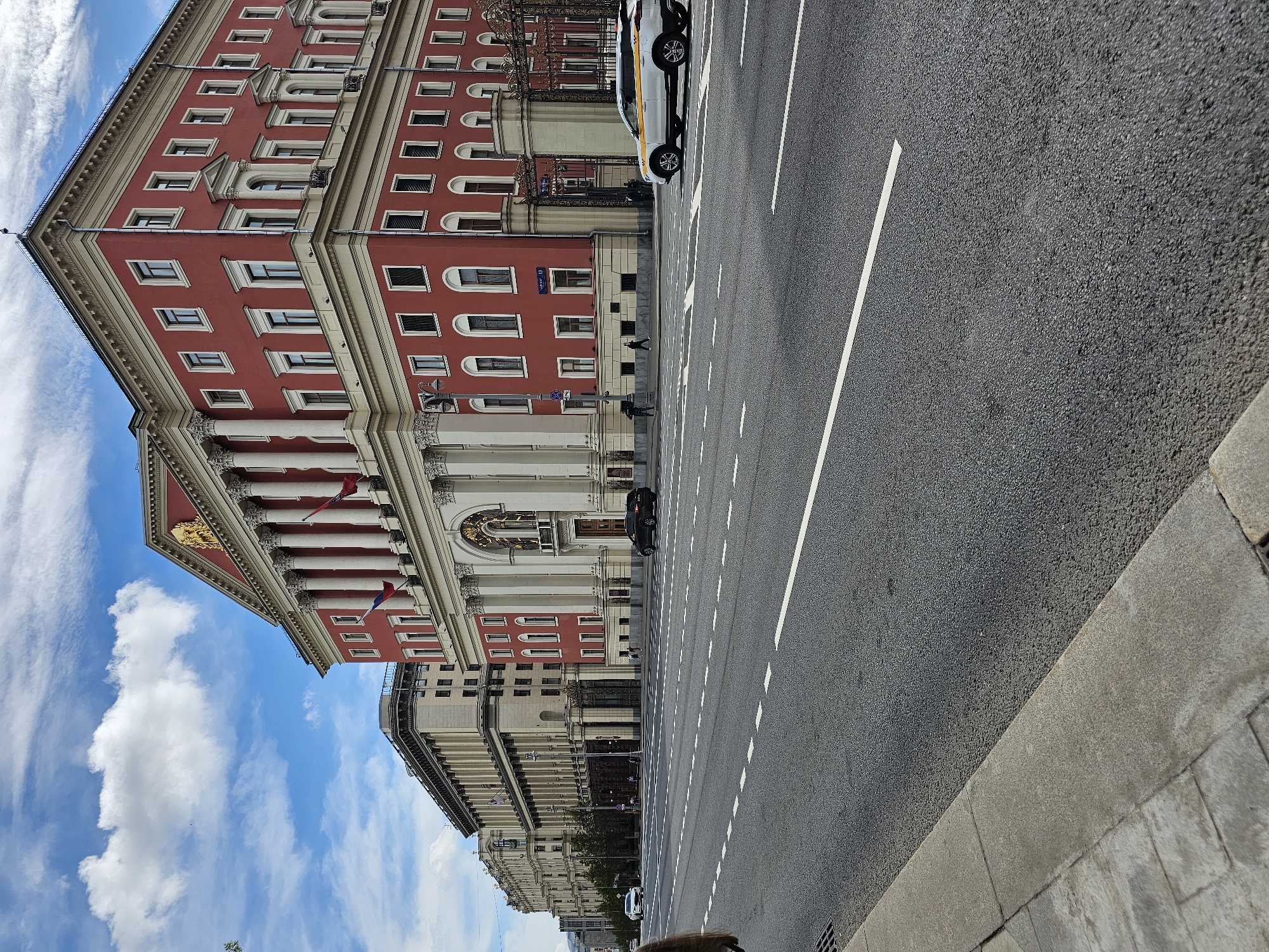 The image captures a view in Moscow, Russia, showcasing the city's architecture and street scene. Prominently featured on the left is a large, multi-story building with a red facade and beige trim, adorned with classical architectural elements such as columns and intricate detailing. A golden emblem or coat of arms is visible on the building's front. The sky above the building is bright blue with scattered white clouds.
To the right of the building, a wide, grey asphalt road stretches into the distance, marked with white lines indicating traffic lanes. Several vehicles are parked along the building's side of the street, including what appears to be a white taxi with yellow accents. Pedestrians can be seen walking on the sidewalk adjacent to the road.
The overall scene conveys a sense of urban activity and the blend of classical architecture with modern city life in Moscow.