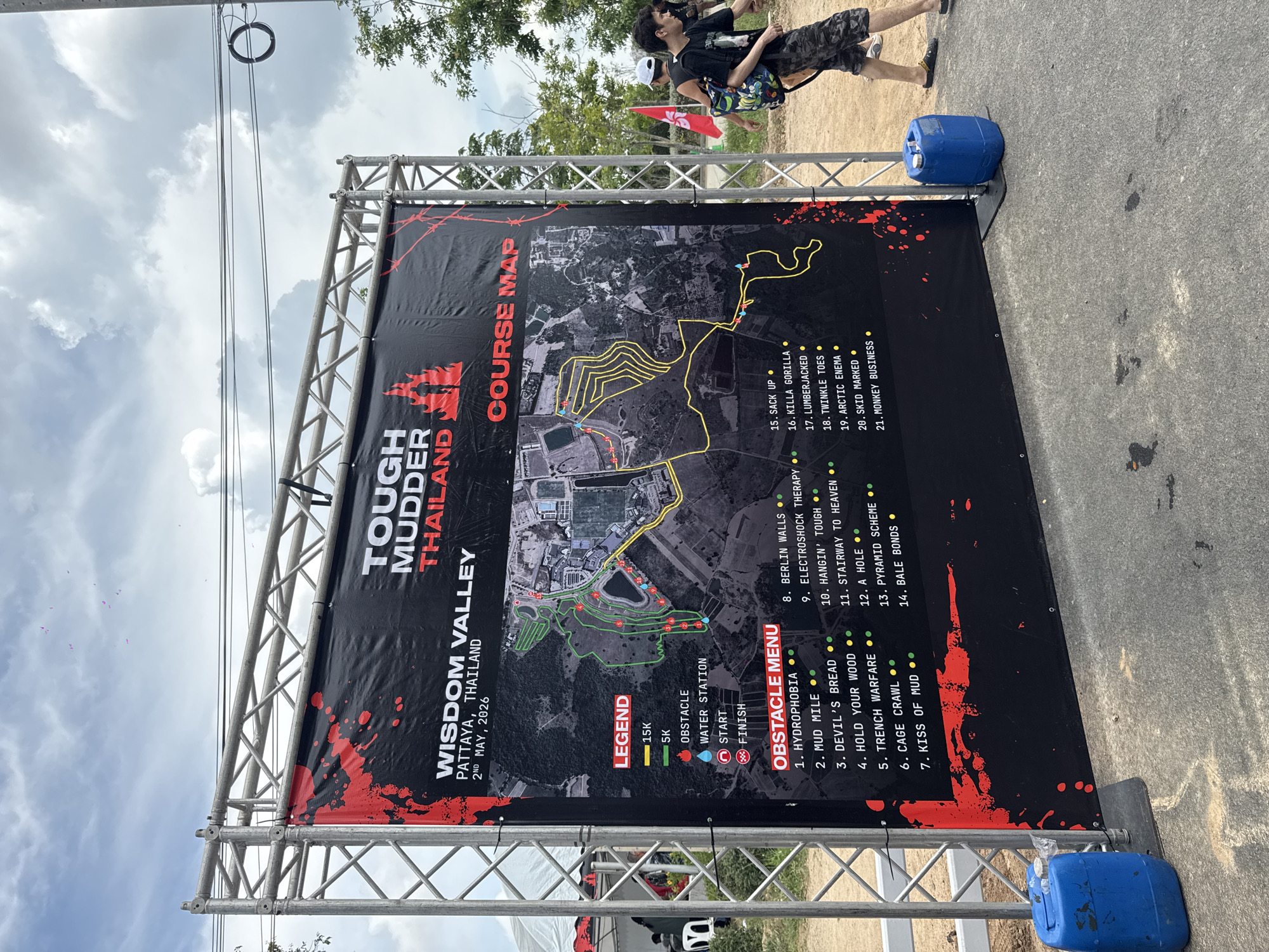 Here is a detailed description of the image:
**Scene:**
The image shows a banner for a "Tough Mudder Thailand" event. The event is to be held at Wisdom Valley in Pattaya, Thailand, on May 2nd, 2026. The banner is held up by a metal frame and secured with blue jugs at the bottom.
**Banner Details:**
The banner displays the event's name, location, and date. A course map is prominently featured on the banner. The map outlines both a 15K and 5K course, marked by yellow and green lines respectively. The legend details that red markers indicate obstacles, blue markers denote water stations, a red circle indicates the start, and the red intertwined rings indicate the finish line. The course map is overlaid on what appears to be a satellite image of the Wisdom Valley area. An "Obstacle Menu" lists the various obstacles along the course, labeled numerically.
**Setting/Environment:**
The banner is set up on a paved road. The background features trees and a cloudy sky.
**People:**
Part of two people can be seen in the upper-right corner of the image. One is wearing a black shirt and camouflage pants and carrying a blue shoulder bag, while the other one is wearing a white hat and black backpack.
**Location:**
The image is most likely taken in the vicinity of Wisdom Valley in Pattaya, within the city of บ้านสำนักตะแบก, Thailand.
**Additional details:**
The background is overcast, suggesting cloudy weather during the photograph.