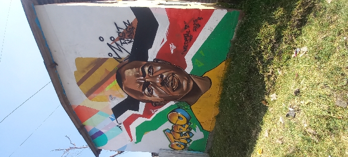 The image displays a graffiti mural covering the majority of a light-colored wall on a single-story building in Nairobi, Kenya. The central element of the mural is a portrait of a dark-skinned male, depicted from the chest up, wearing a yellow t-shirt and looking directly forward with a serious expression. The background of the portrait features the colors of the Kenyan flag (black, red, green, with white fimbriations). To the left of the figure, "OTERO" is rendered in stylized yellow, blue, and black graffiti. To the right, "FAST KINGZ" is visible in black, and below it, "MR. DETAIL 7" and "2025" are inscribed, accompanied by a small white paper airplane symbol. The upper left corner of the mural contains abstract geometric designs in various colors. The building has a dark-edged roof with some blue trim visible. The ground in the foreground is covered with green grass and scattered debris. The sky above is clear and bright blue, with faint power lines visible.