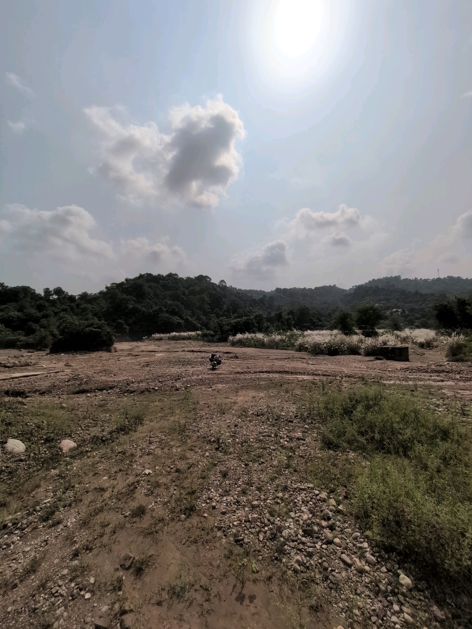 A wide-angle, low-angle shot captures a dry riverbed in the city of Jaanta, India. The terrain is predominantly rocky and sandy, with sparse vegetation. A lone motorcycle is parked in the middle distance, its rider unseen. In the background, a dense forest covers rolling hills. The sky is partly cloudy with a bright sun visible, casting a hazy glow. The overall scene suggests a rural, natural landscape.