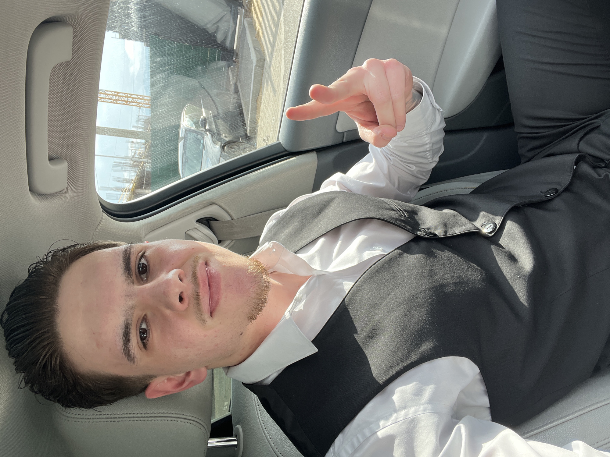 A young adult male is depicted seated in the front passenger seat of a vehicle with a light-colored interior. He possesses dark, slicked-back hair, a mustache, and a goatee. He is dressed in a white long-sleeved collared shirt, a black vest, and black trousers. His right hand is raised towards the viewer, forming a "finger gun" gesture with his index and middle fingers extended. He is looking forward with a slight smile. Strong sunlight illuminates the scene from the right, casting distinct shadows inside the vehicle.
Through the vehicle's passenger window, an outdoor environment is visible. A construction site occupies the foreground, characterized by multiple vertical rebar structures, a prominent yellow lattice boom crane, and a tall green mesh construction barrier. Behind the barrier, a dark-colored sedan is parked on a paved surface. In the distant background, a large mountain range, identifiable as Vitosha Mountain, is visible under a clear to partly cloudy sky. The combination of active urban construction and the distinctive Vitosha Mountain range places the scene within the city of Sofia, Bulgaria.