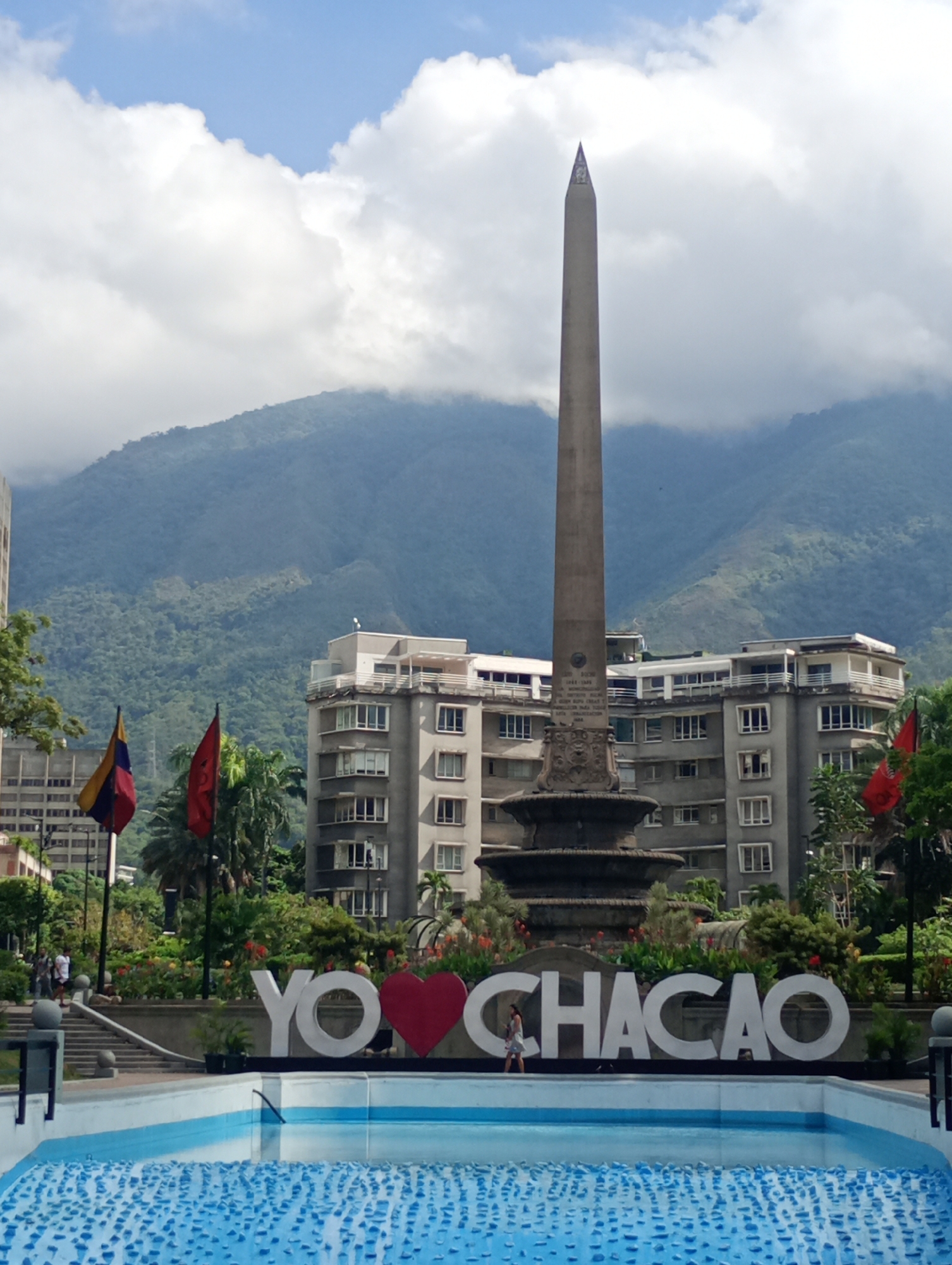 The image depicts Plaza Francia in Caracas, Venezuela. In the foreground is a light blue water feature or fountain. Behind the fountain is a large white sign reading "YO <3 CHACAO," with a red heart symbol replacing the word "love." Behind the sign stands the plaza's central monument, an obelisk-like structure with decorative elements at its base. Buildings of varying heights and architectural styles are visible behind the monument, and beyond them, a backdrop of green mountains partially obscured by clouds. Flags of Venezuela and possibly other entities are present on poles to the left and right of the monument. A few people are visible in the background, walking around the plaza.