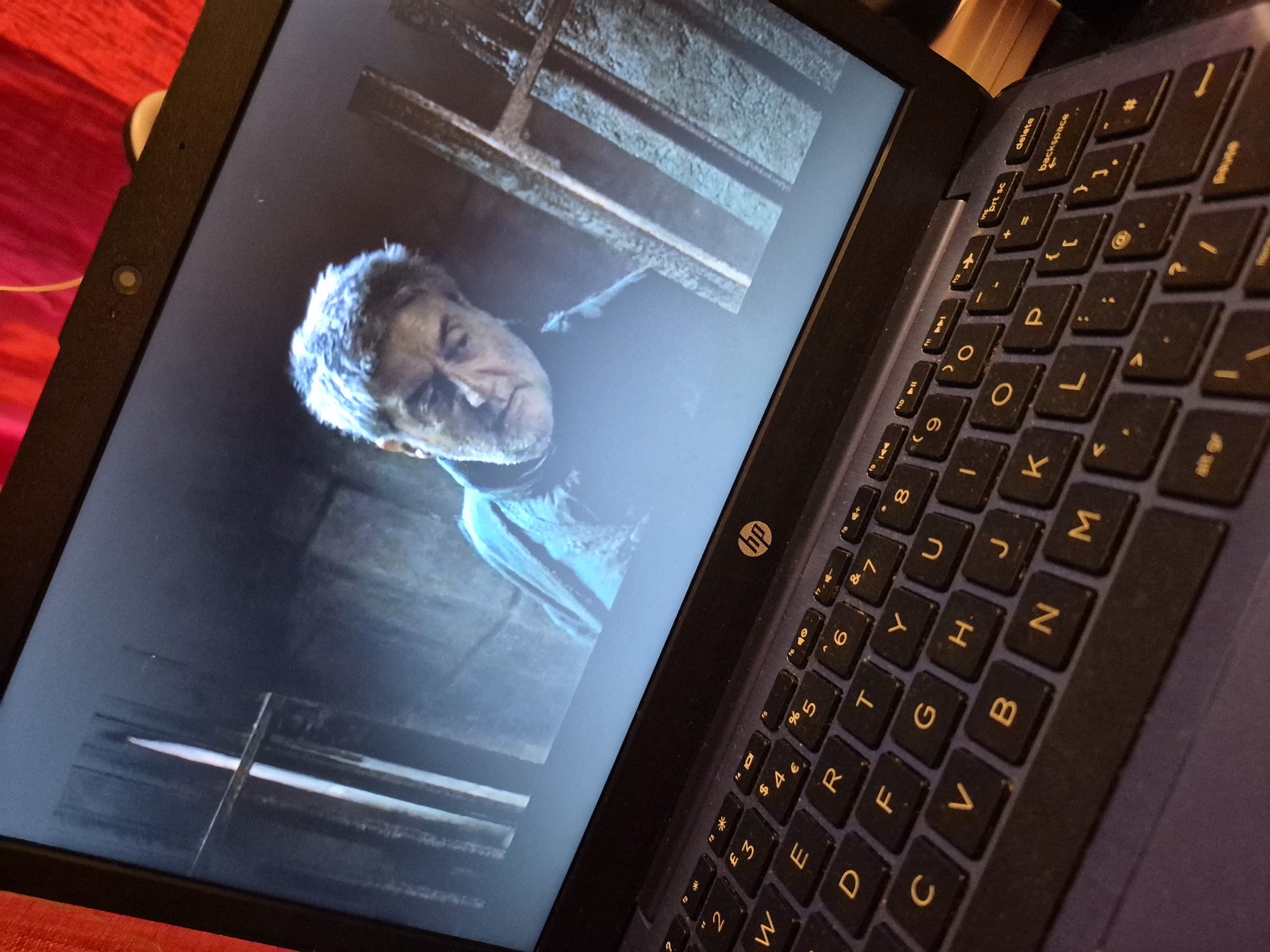 A close-up, high-angle, vertical shot shows a dark blue laptop open and displaying a scene from a movie or show. The screen shows the upper body of a man with graying hair and a beard, wearing a dark blue garment. He appears to be in a dimly lit, possibly industrial or prison-like setting, with bars or vertical structures visible in the background. The laptop is placed on a dark surface, and a red fabric is visible in the upper left corner of the frame, possibly indicating the environment where the laptop is situated. The keyboard of the laptop is clearly visible in the foreground, with keys illuminated by the screen's glow. The HP logo is visible on the laptop. There is no discernible connection to the city of Sandwell, United Kingdom in this image.