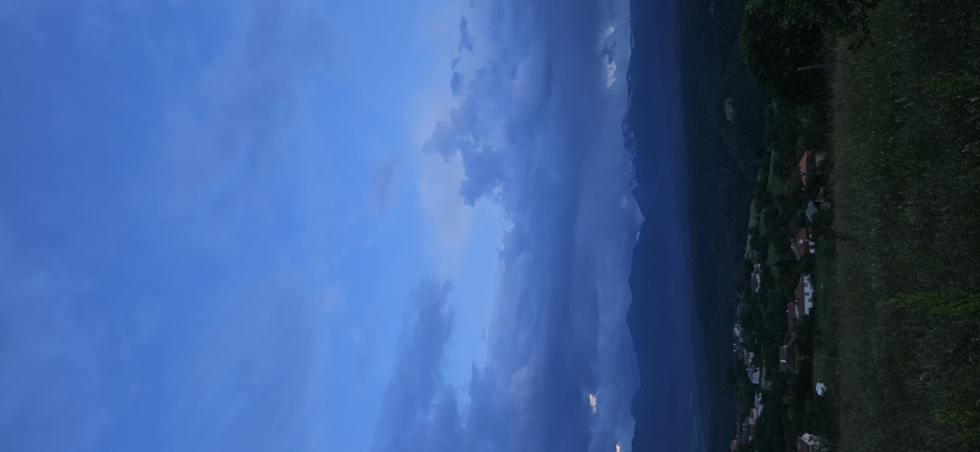 The image displays a sprawling mountainous landscape under a dramatic twilight sky, indicative of either late evening or early morning. The sky dominates the upper and left portions, showcasing a deep blue hue that shifts from a lighter, clearer blue to darker, more intense shades where voluminous clouds gather, particularly on the right side. These clouds create a sense of depth and a slightly overcast or moody atmosphere.
A prominent mountain range stretches across the horizon in the mid-ground, appearing dark blue and imposing against the sky, emphasizing its grand scale. Below the mountains, the terrain is characterized by lush green hills or fields. Towards the lower right, a rural village is nestled within this verdant landscape. Several houses with distinct red or orange tiled roofs and lighter-colored walls are visible, interspersed among trees and other vegetation.
No specific activities or people are discernible in the static scene. The overall lighting is dim and diffused, lending a serene yet somewhat solemn or vast quality to the environment. There is no visible text. The location depicted is Gutsal, Bulgaria.